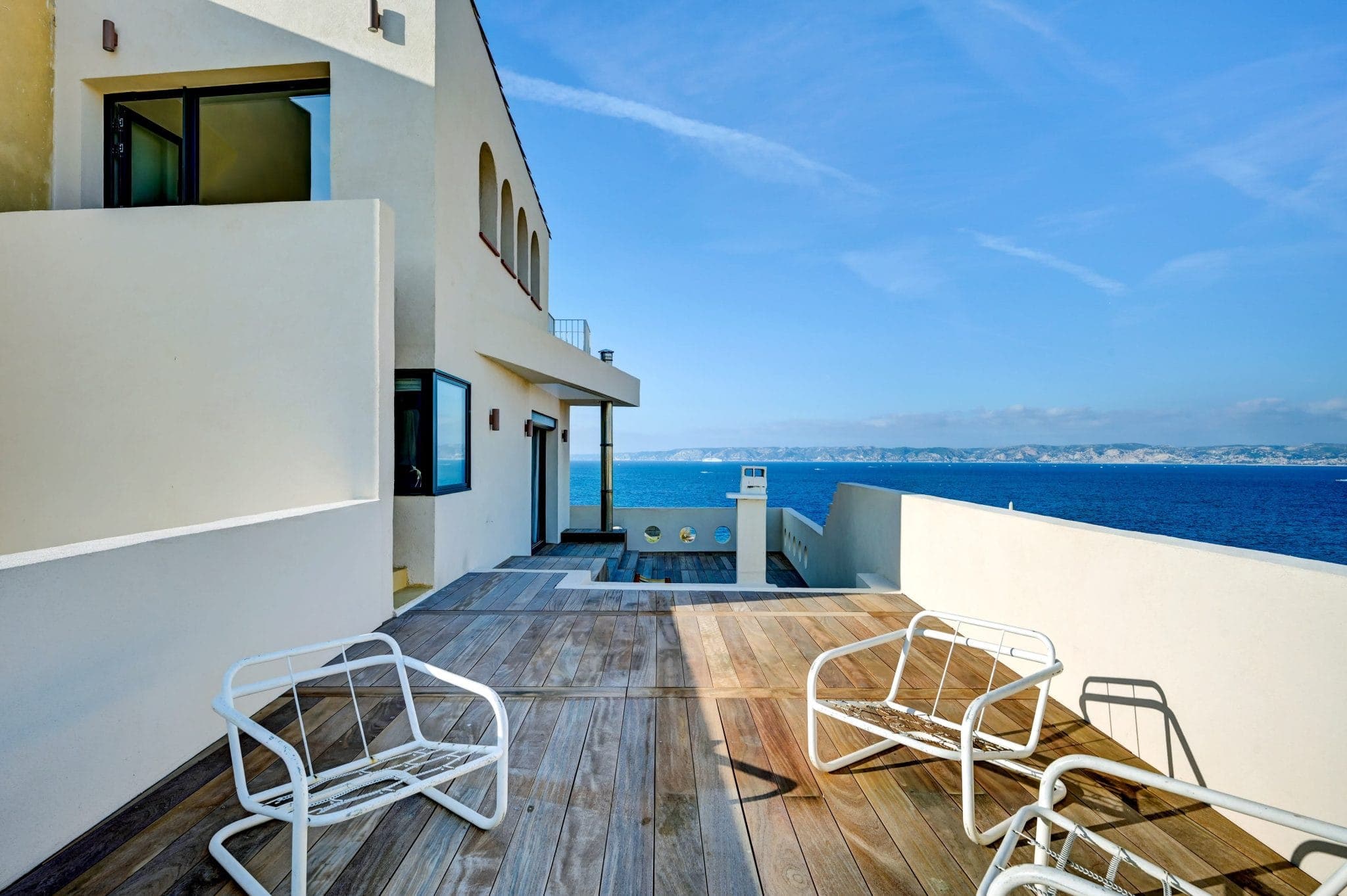 Une terrasse moderne en bord de mer avec des chaises blanches sur une terrasse en bois surplombe un vaste océan bleu. Le bâtiment dispose de grandes fenêtres et d'ouvertures cintrées, avec un ciel clair et un littoral lointain en arrière-plan.
