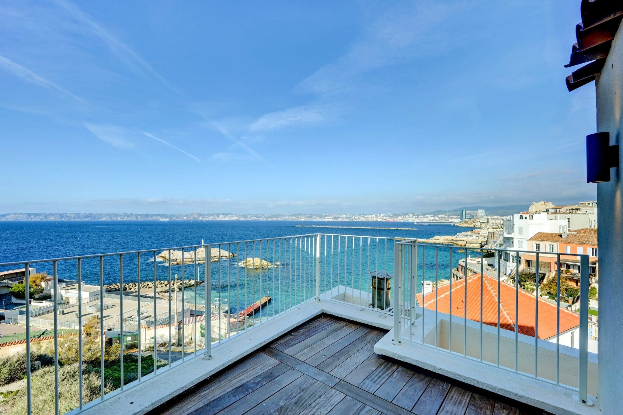 Vue panoramique d'une ville côtière depuis un balcon avec terrasse en bois. L'image montre un ciel bleu vif, une mer limpide et des bâtiments aux toits rouges. Une balustrade en métal entoure la terrasse, améliorant la vue panoramique sur le front de mer et le paysage urbain.