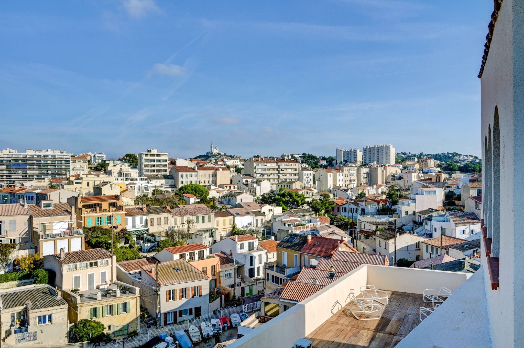 Vue panoramique d'une ville côtière avec un mélange de bâtiments modernes et traditionnels sous un ciel bleu clair. Le premier plan montre une terrasse sur le toit avec des meubles d'extérieur, tandis que l'arrière-plan présente des structures urbaines densément peuplées et des collines lointaines.