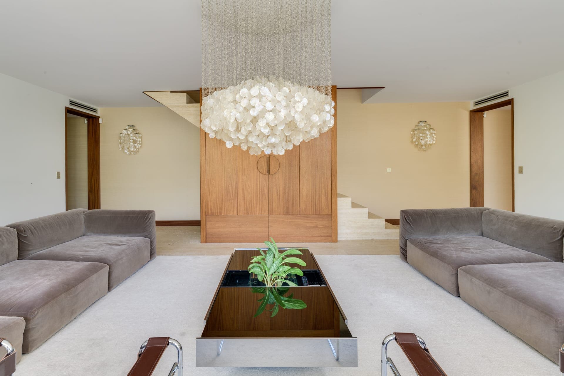 Un salon moderne est doté de canapés gris, d'une table basse en verre avec une plante et d'un grand lustre complexe. Des boiseries et un escalier sont à l'arrière-plan. L'espace est minimaliste et aéré.