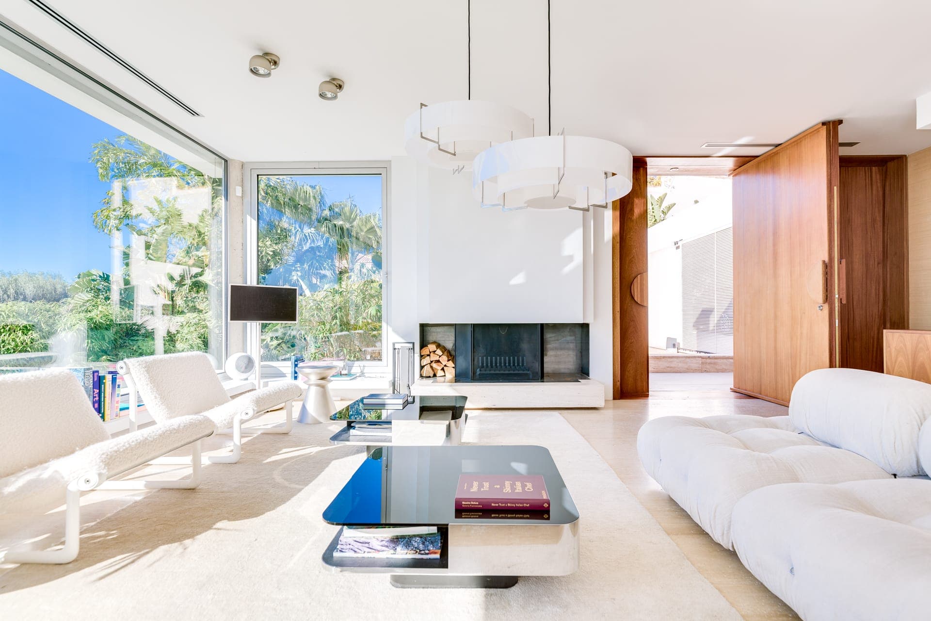 Un salon lumineux et moderne avec de grandes fenêtres offrant une vue sur le jardin. Il comprend des chaises longues blanches, un canapé moelleux, une table basse contemporaine avec des livres et une cheminée minimaliste. Des suspensions sont suspendues au plafond et une porte est entrouverte.