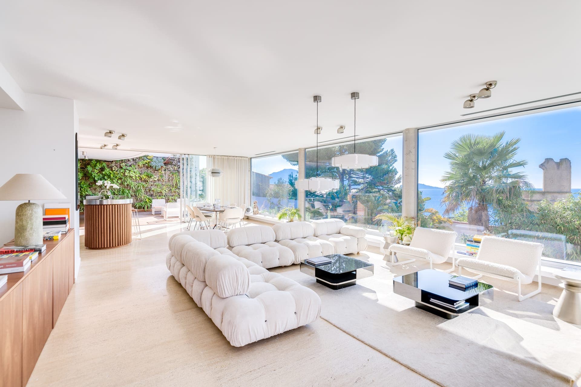 Un salon lumineux et moderne avec de grandes fenêtres offrant une vue panoramique sur les palmiers et les montagnes. La pièce comprend des canapés blancs, des tables noires, des luminaires suspendus et un buffet en bois. Au fond, il y a un coin repas avec de la verdure.