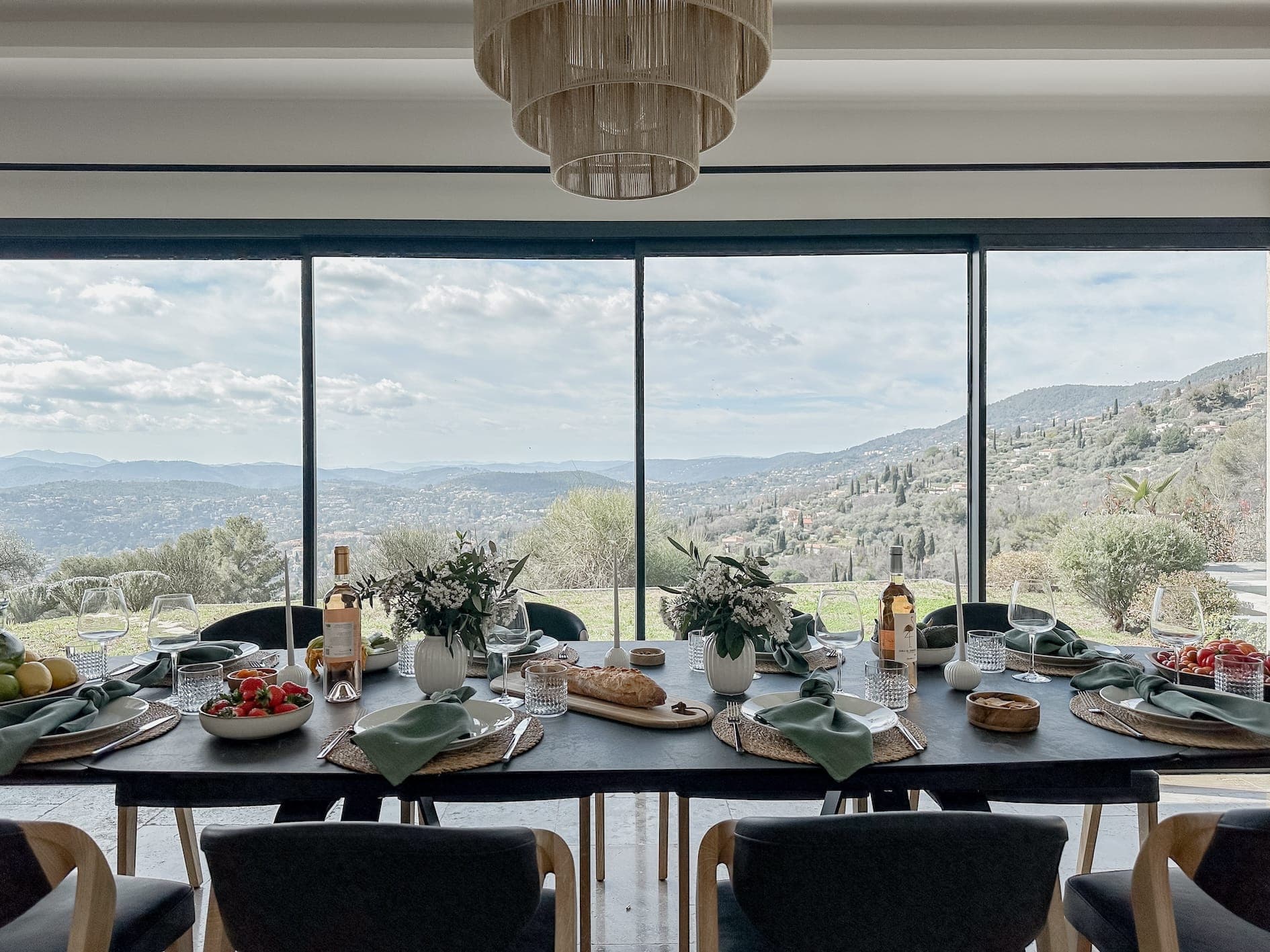 A dining table set for a meal with plates, wine, bread, and fruits. The table is positioned in front of large windows, offering a scenic view of a picturesque valley and mountains under a partly cloudy sky. A chandelier hangs above the table.