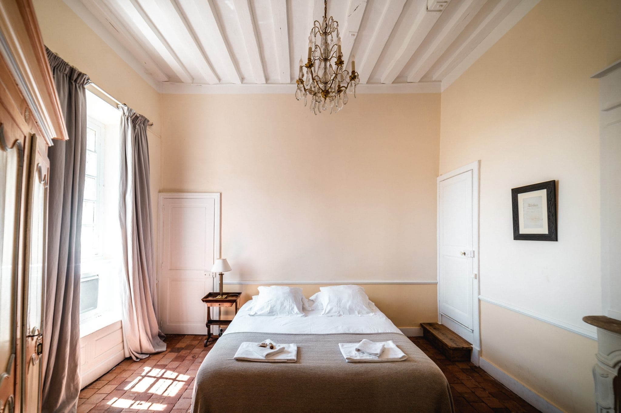 A comfortable room with a well-made double bed, two white pillows and fresh towels. Soft lighting from a chandelier illuminates the room. Wooden floors, a large window with gray curtains and minimalist decor create a welcoming ambience.