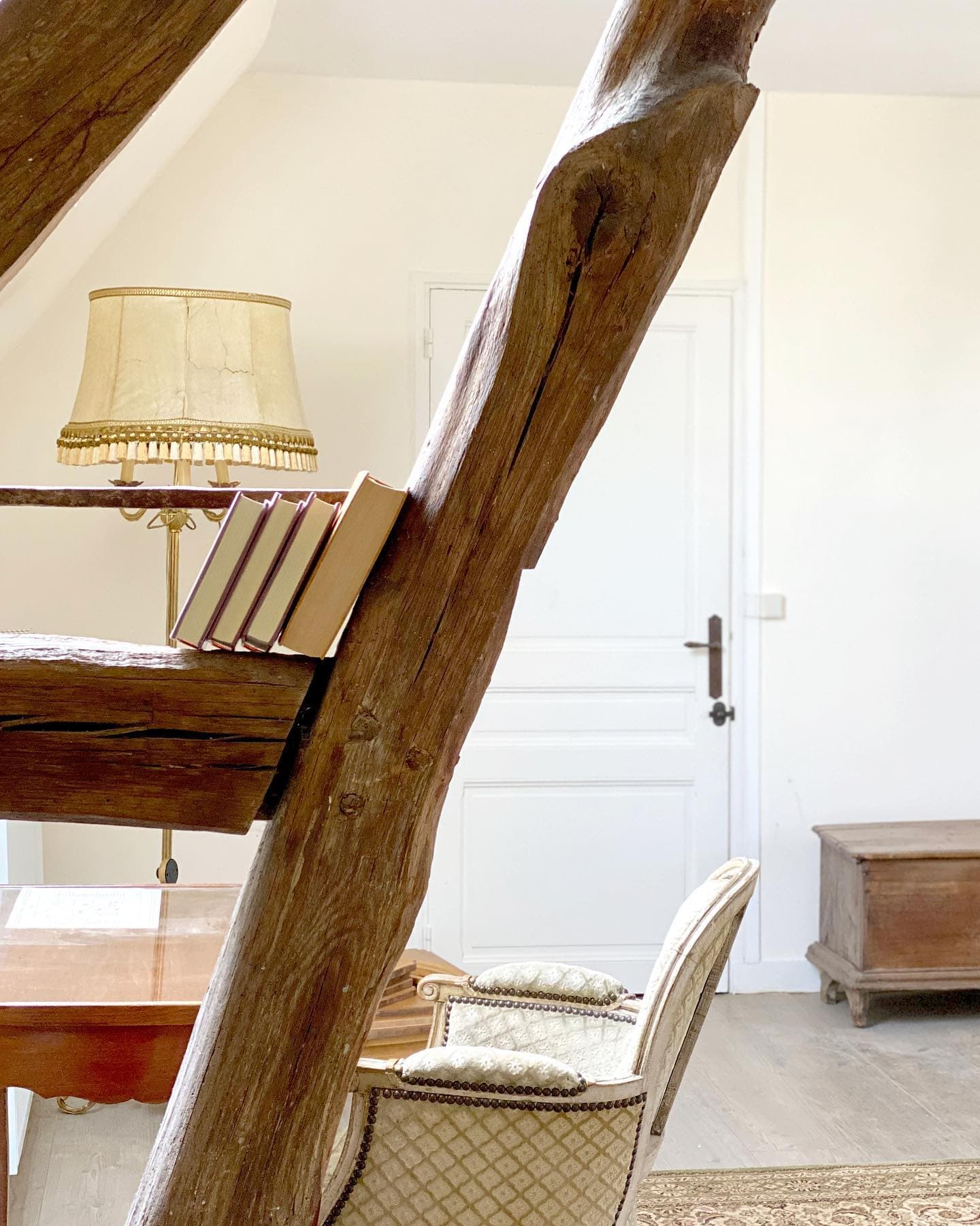 A warm room with a rustic wooden staircase, a vintage chair and a wooden chest. Books are lined up on the stairs and a decorative lamp sits on a table next to a white door. The atmosphere is warm and welcoming.