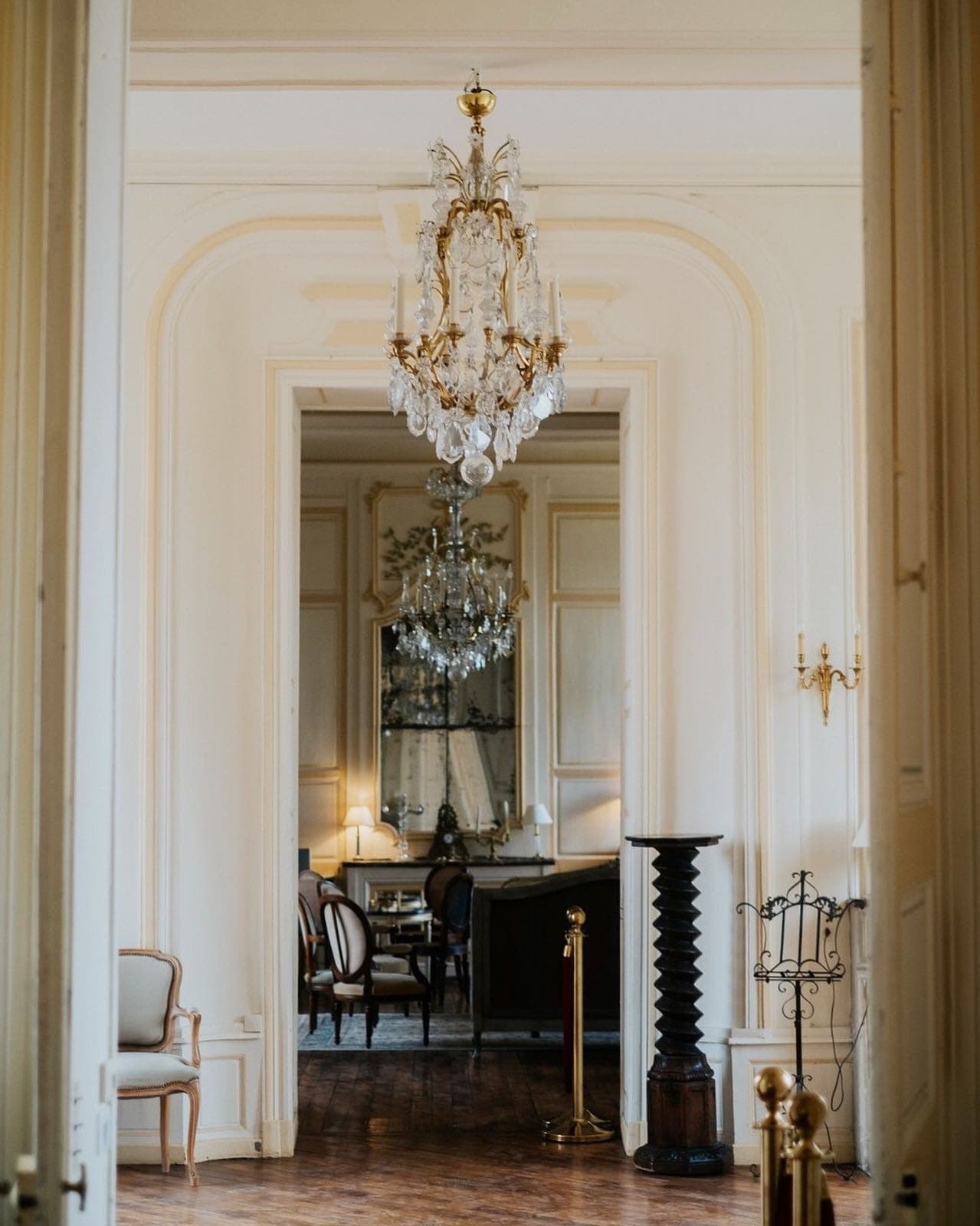 A luxurious interior with elegant chandeliers, richly detailed moldings, antique furniture and polished wood floors. The hallway leads to a majestic room with a large mirror and opulent decor, evoking a classic, refined atmosphere.