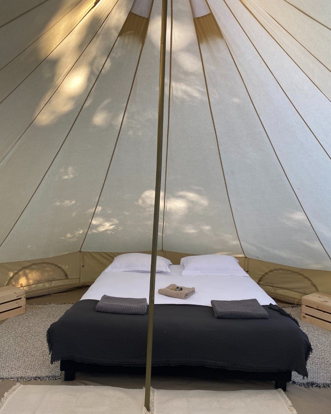 Interior view of a glamping tent featuring a carefully made bed with white pillows, a dark blanket and folded towels. Wooden crates serve as bedside tables, and soft natural light filters through the tent fabric.