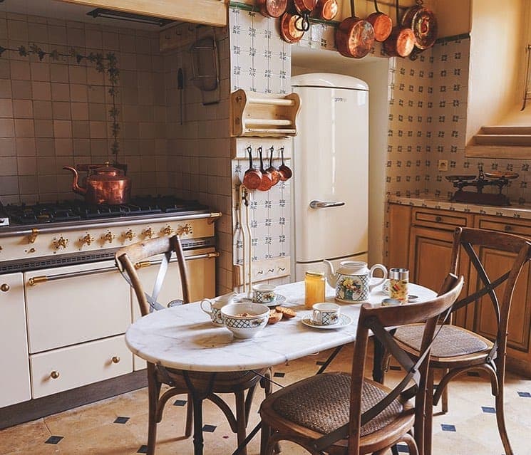 A vintage-style kitchen with a retro white refrigerator, a gas stove with a copper pot and a small oval table with teacups and a teapot. Copper pots and pans hang over the stove, giving the room a warm, cozy feel.