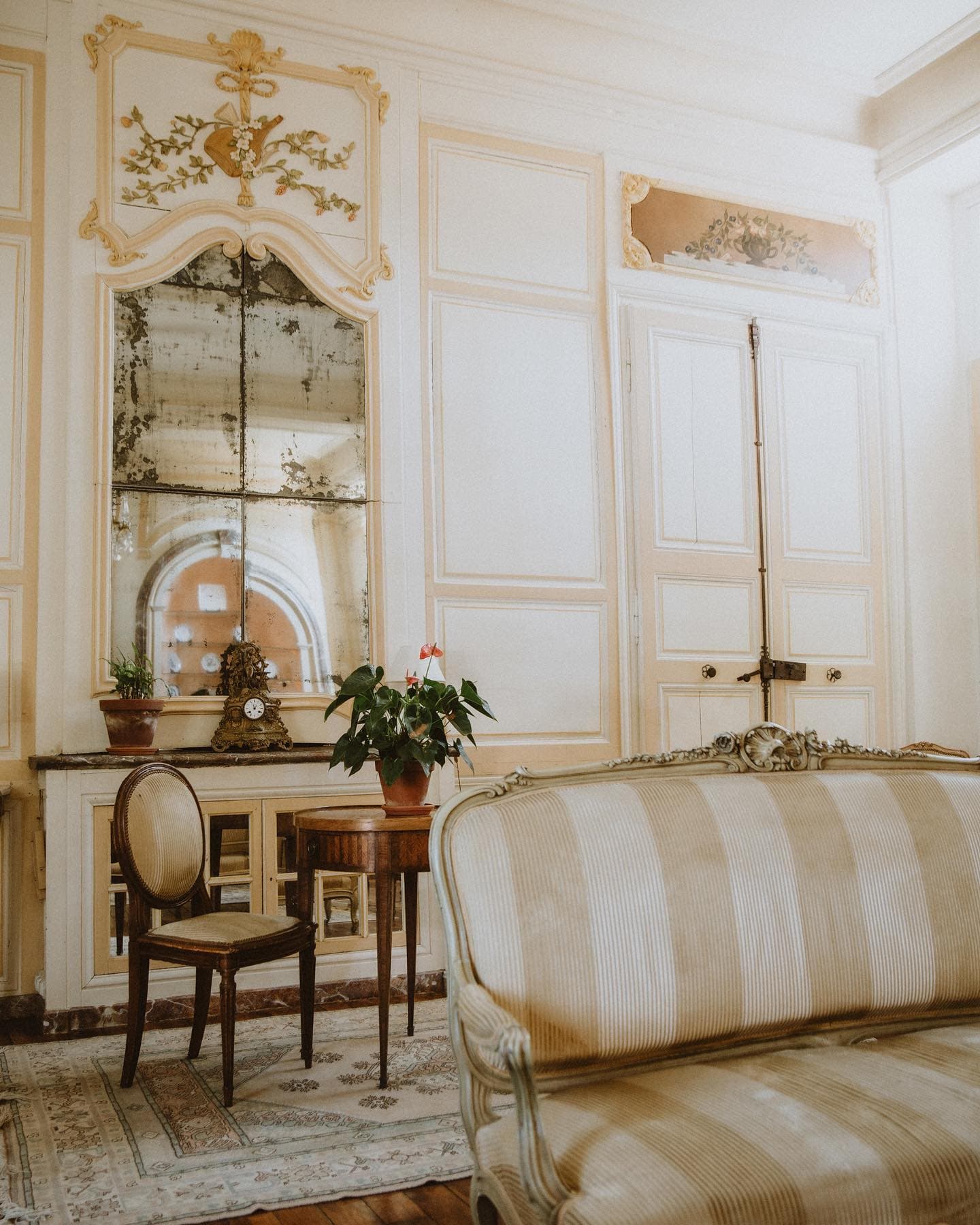 Elegant vintage room with ornate mirror, decorative moldings and floral arrangement. A wooden chair and rustic armoire accessorize the space. A striped, upholstered sofa takes pride of place in the foreground, contributing to the classic, sophisticated ambience.