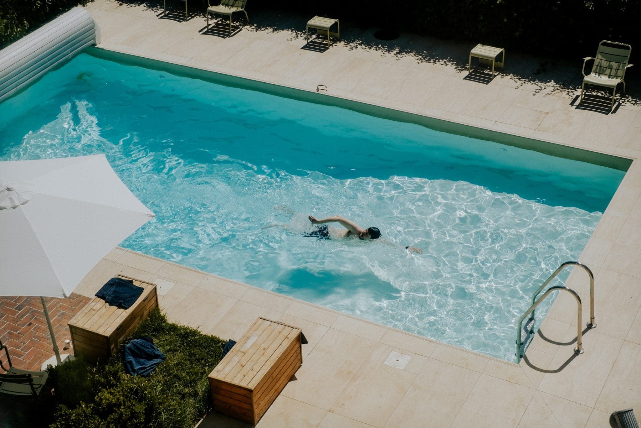 One person is swimming in a rectangular outdoor pool surrounded by a beige terrace. There are sun loungers and a white parasol at one end. The water is clear and blue, the light reflecting off the surface.