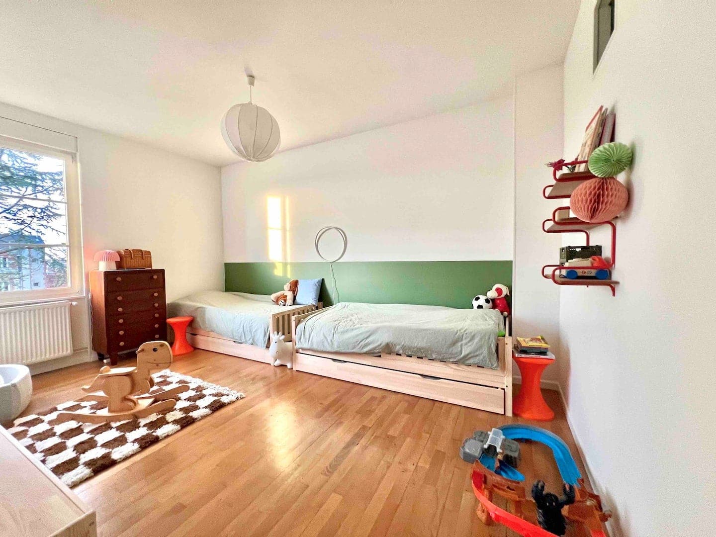 A bright nursery with two single beds and green-accented walls. A wooden rocking horse and a soft rug lie on the floor. Shelves with toys and books are fixed to the wall, and a chest of drawers stands by the window, letting in the sunlight.