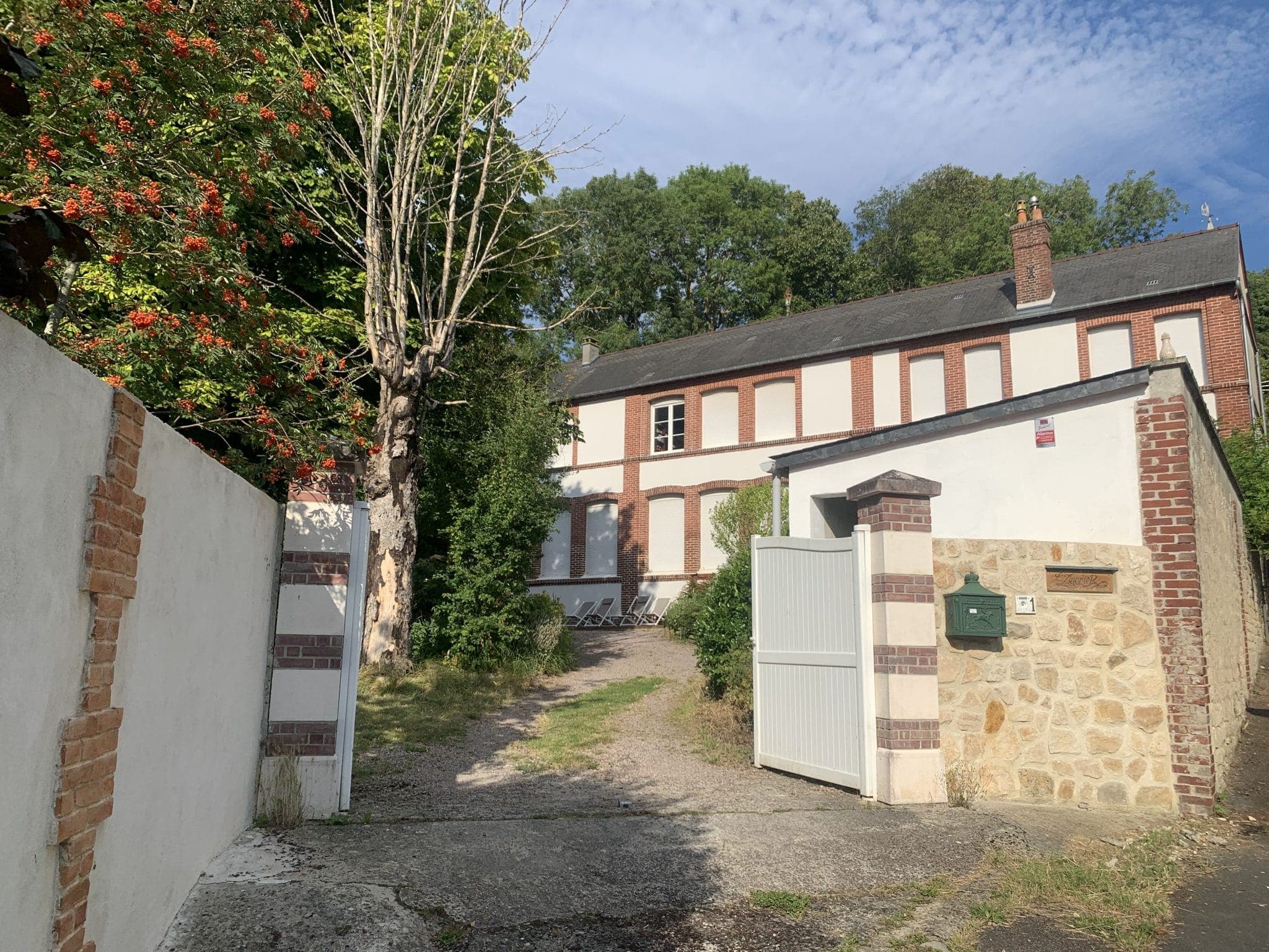 Behind an open gate with brick pillars, a two-storey brick house with white shutters comes into view. A tree with red berries stands to the left, and grass lines the gravel driveway leading to the house. The sky is partly cloudy.