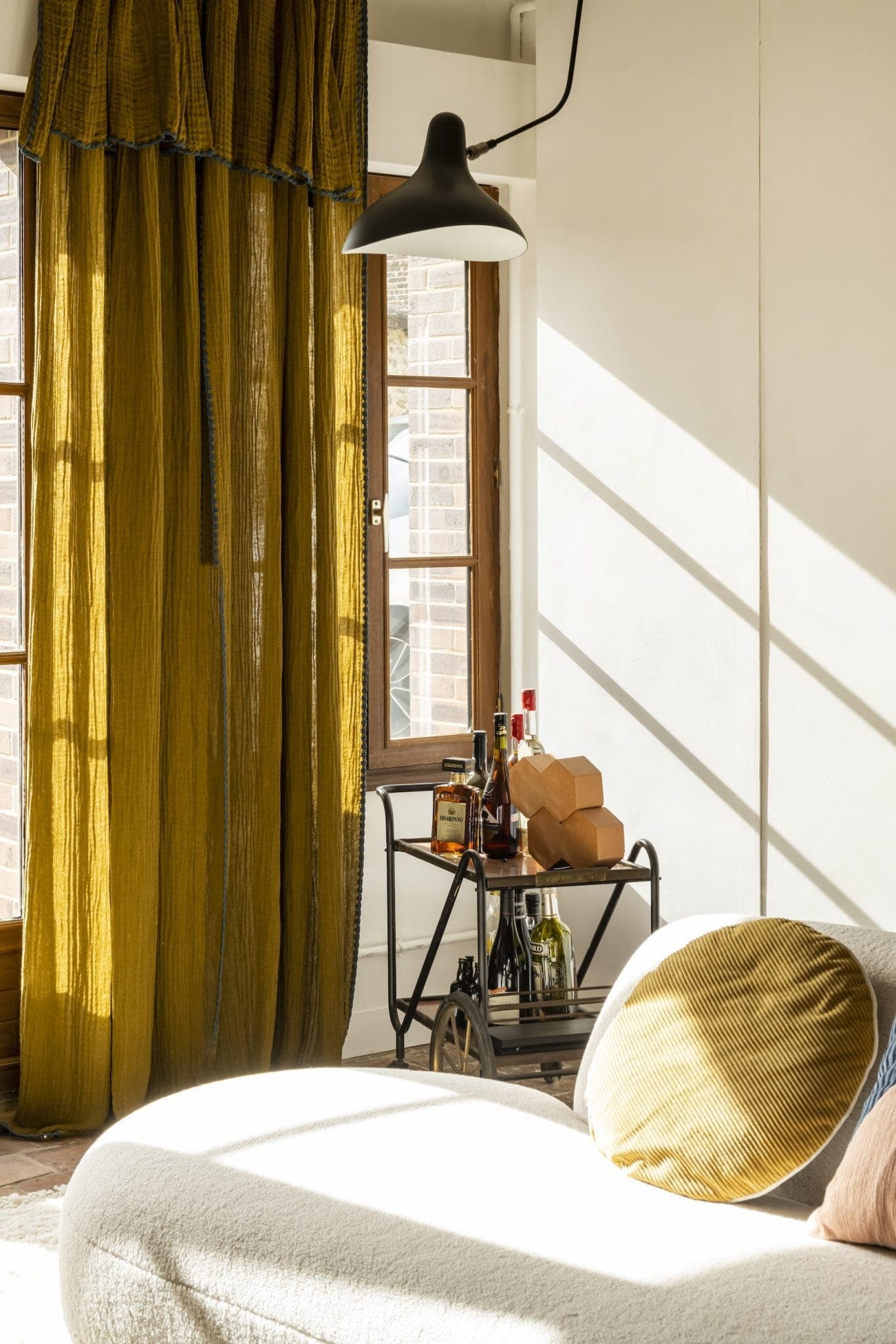 Un coin salon confortable avec des rideaux jaunes et une lumière du soleil qui entre à flot. Un chariot de bar accueille des bouteilles et des verres. Un canapé rond blanc avec un coussin moutarde est au premier plan. Une suspension noire est suspendue au-dessus.