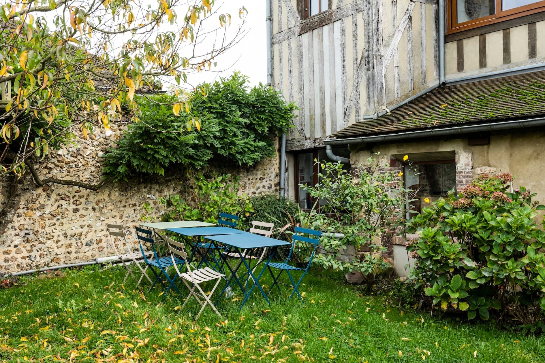 Un jardin pittoresque avec une table en métal bleu entourée de chaises dépareillées sur de l'herbe verte. Le jardin est bordé de murs en pierre et de plantes luxuriantes, et une maison rustique aux murs en bois rayés est visible en arrière-plan.