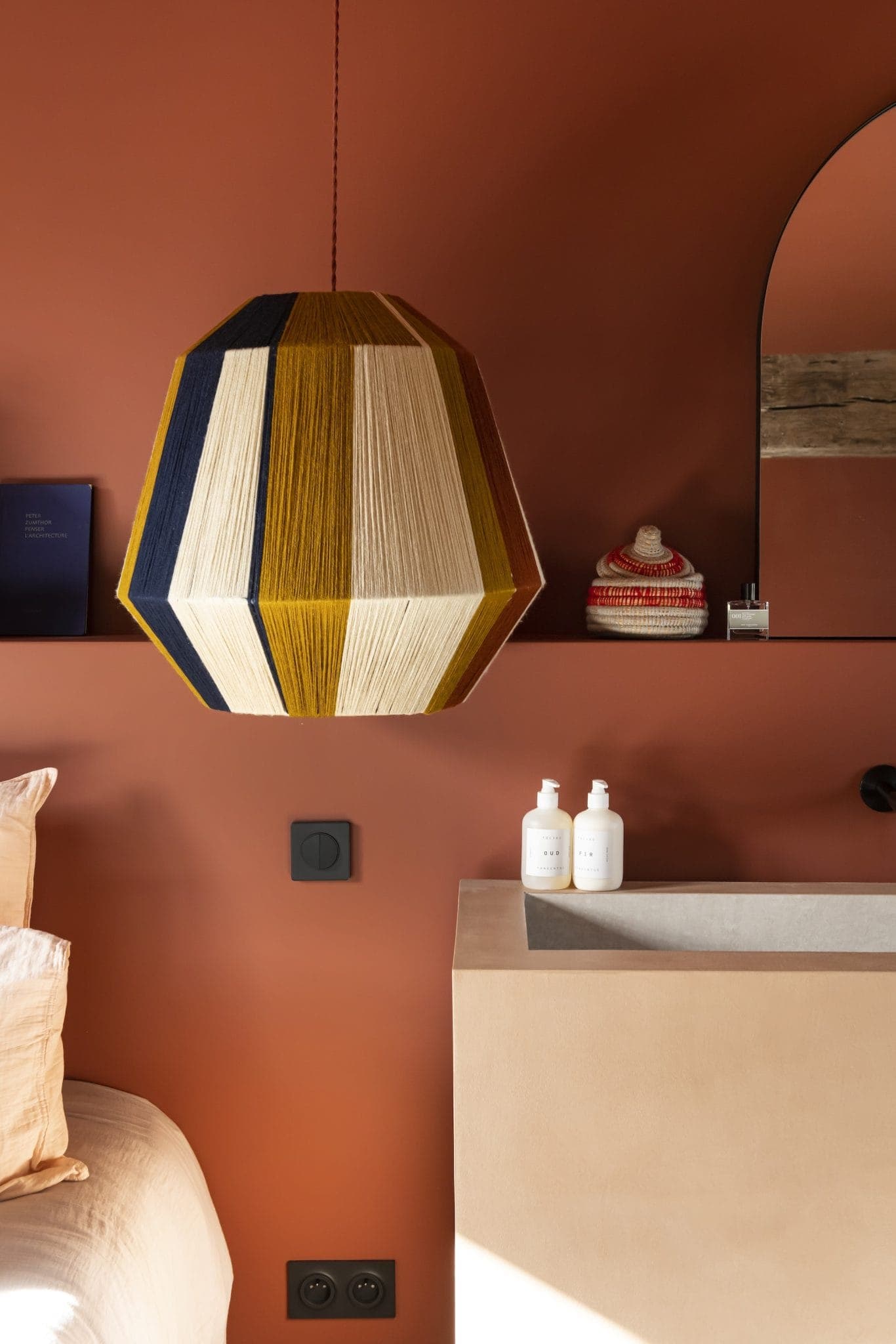 Une lampe à suspension géométrique à rayures multicolores est suspendue au-dessus d'un comptoir de salle de bains minimaliste avec deux bouteilles de lotion. Un miroir reflète un bonnet tricoté et un livre repose sur une étagère. Les murs sont d'une couleur terre cuite chaleureuse.