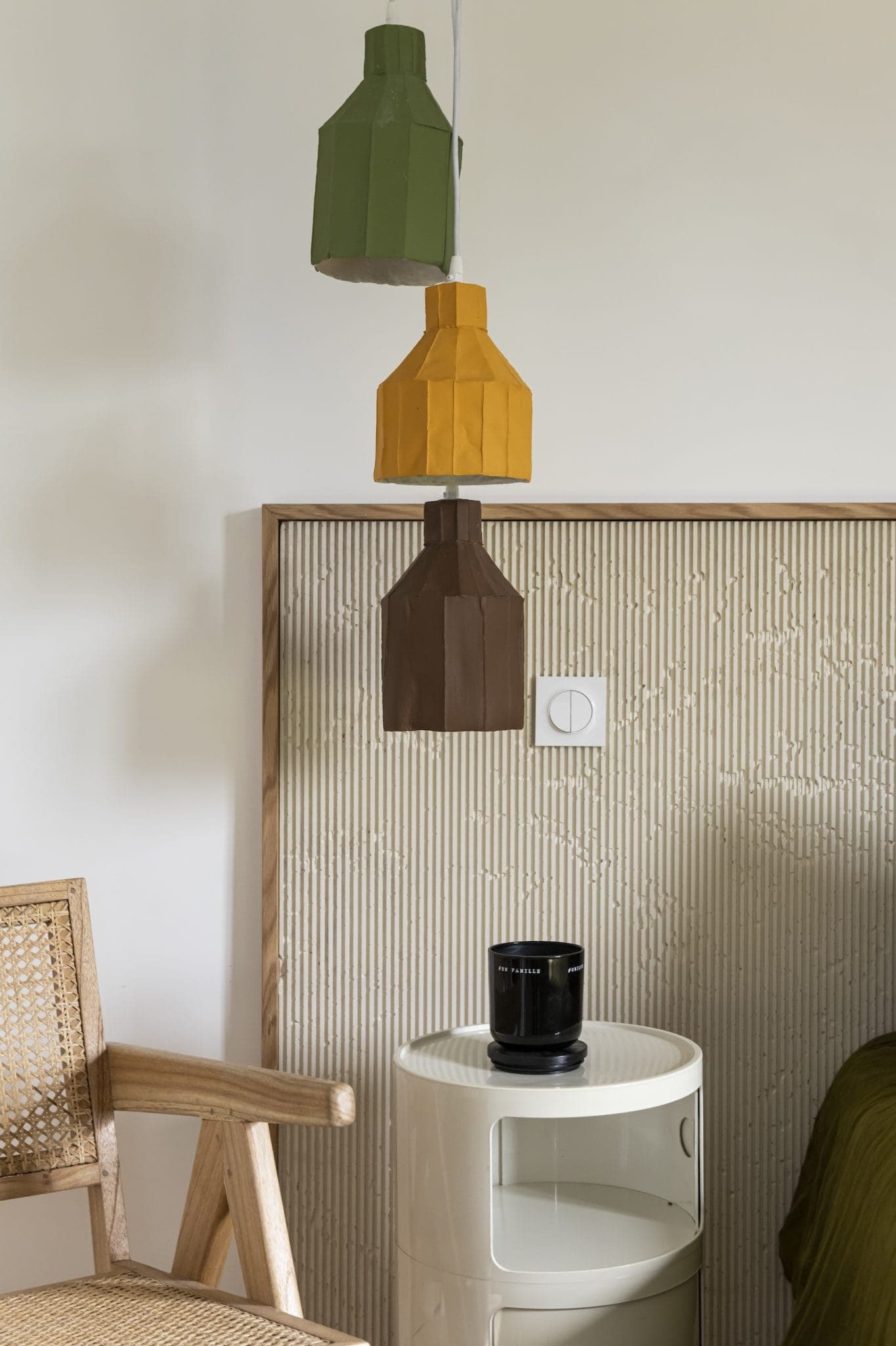 Une chambre moderne avec une chaise en bois, une table d'appoint ronde avec une bougie noire et une tête de lit texturée. Un trio de lampes suspendues géométriques vertes, jaunes et brunes sont suspendues au-dessus de la table. Des murs et une literie neutres complètent le décor minimaliste.