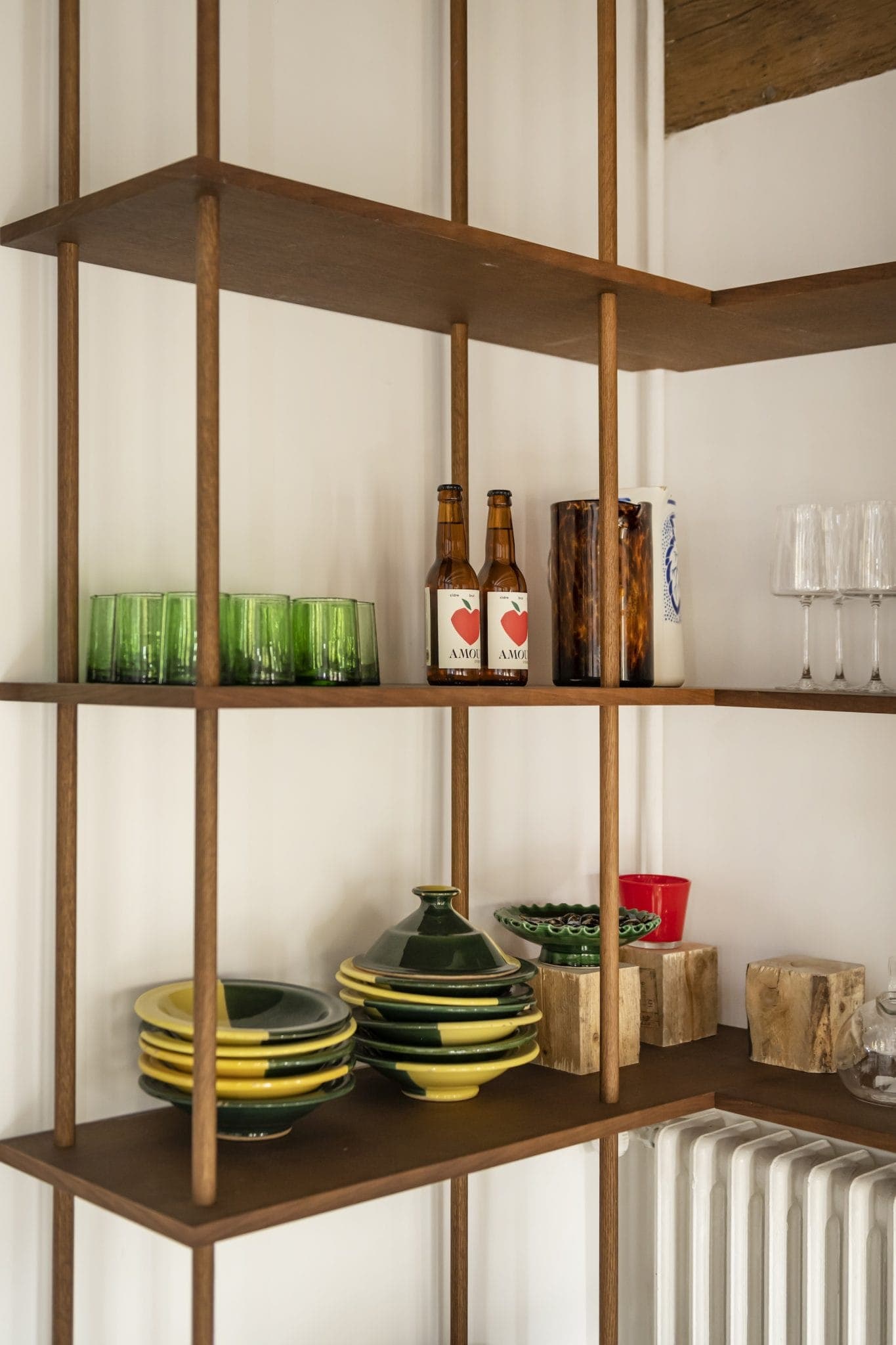Une étagère en bois accueille des verres verts, des assiettes, deux bouteilles, des bols en céramique et de la verrerie. Le design minimaliste de l'étagère présente des supports verticaux et un espace de rangement ouvert, créant un affichage organisé contre un mur neutre.