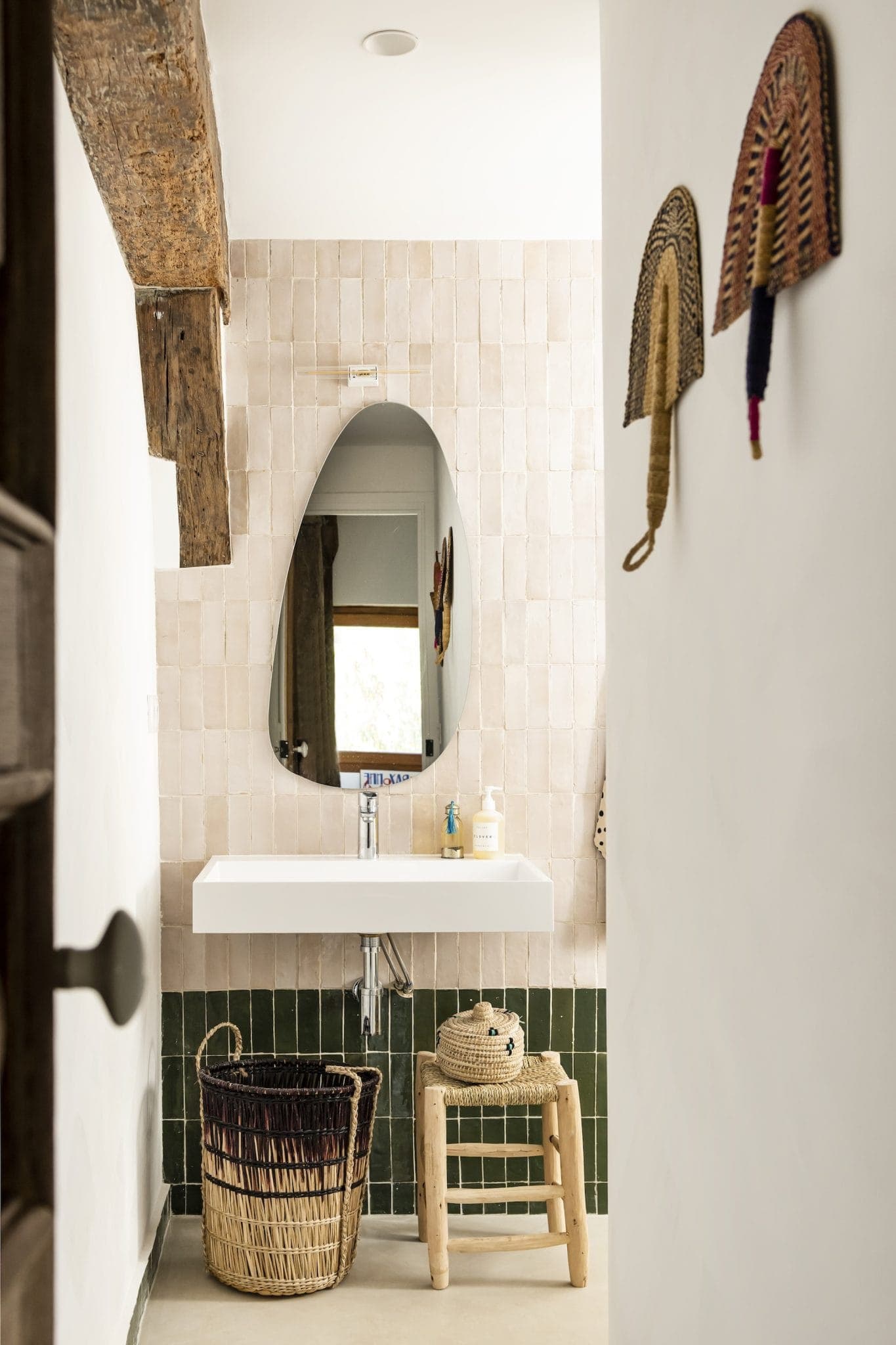 Une salle de bains rustique est dotée d'un miroir ovale au-dessus d'un lavabo rectangulaire, de carrelage beige clair et d'une poutre en bois au-dessus. Un panier tressé et un tabouret en bois sont placés en dessous, et des tentures murales décoratives sont visibles sur le côté.