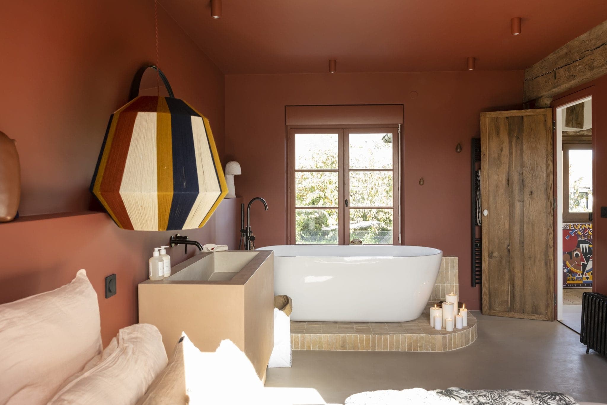 Une chambre confortable et moderne aux tons terreux dispose d'une baignoire blanche autoportante sous une fenêtre. Une grande suspension géométrique est suspendue au plafond. Une porte en bois rustique est entrouverte et des bougies allumées sont placées près de la baignoire.