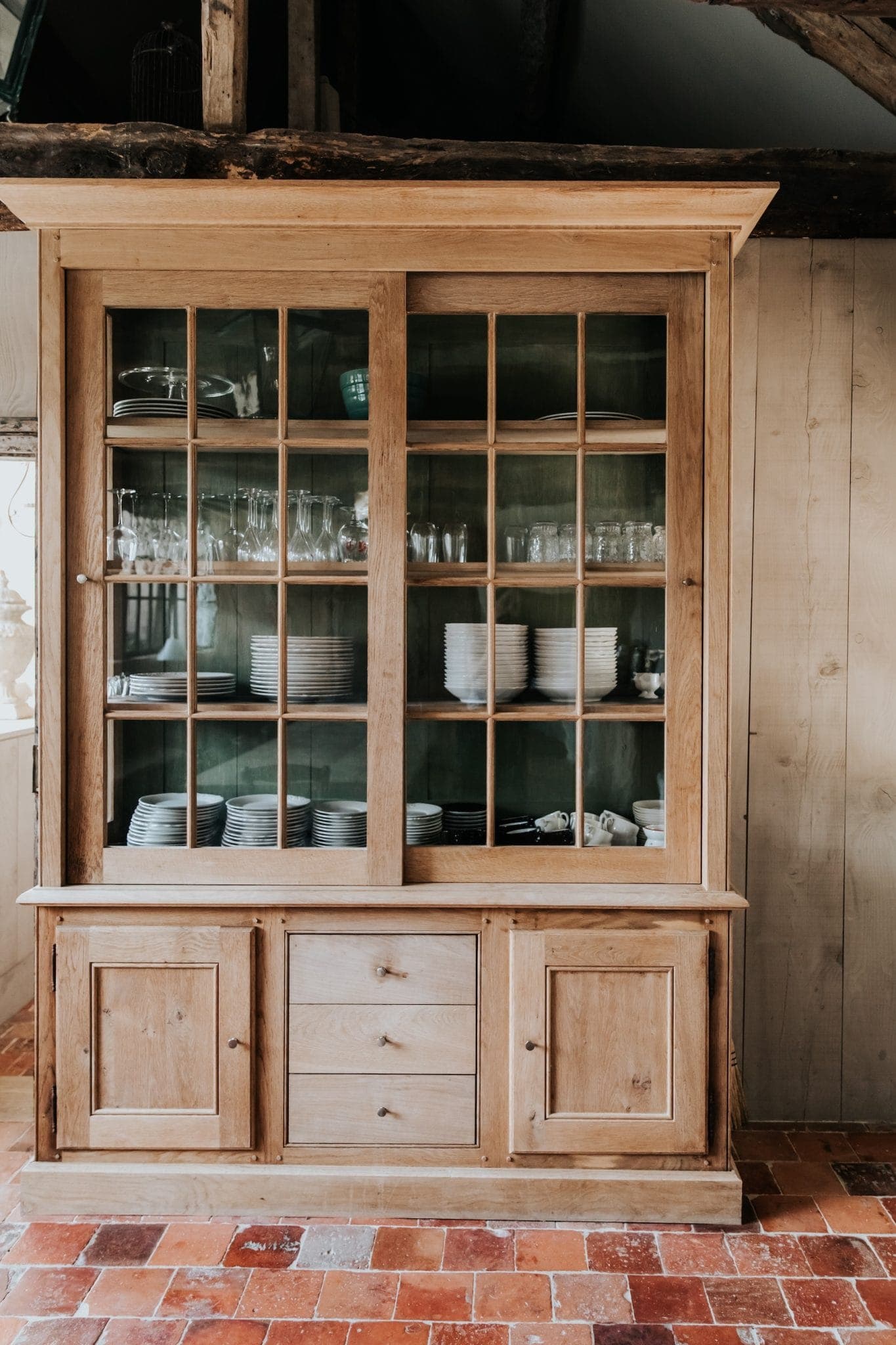 A wooden showcase with glass doors featuring carefully arranged plates, bowls and glasses. It stands against a wooden wall on a tiled floor, blending rustic and warm design elements.