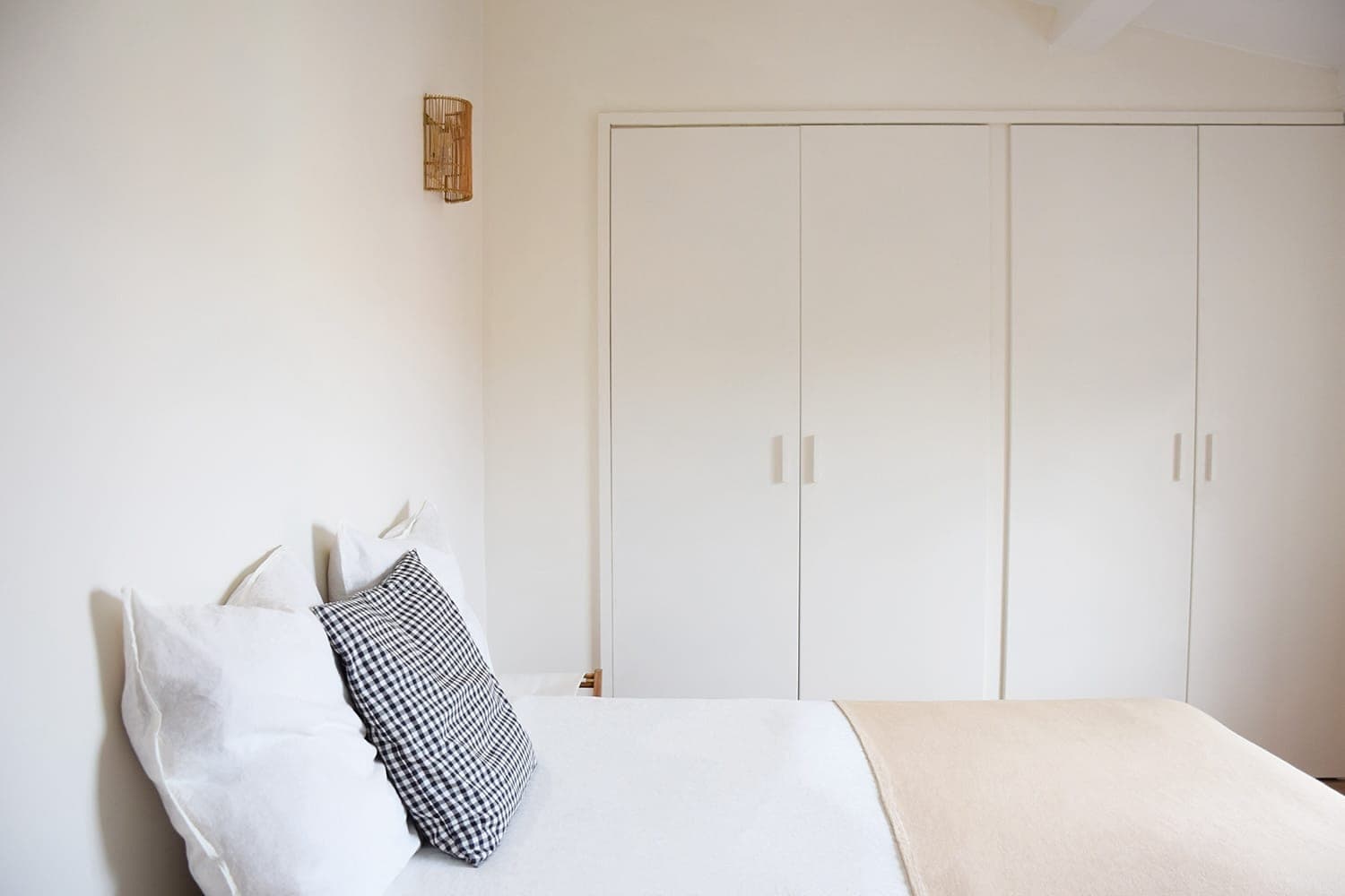 Minimalist room with a well-made bed and white and beige sheets. A plaid pillow adds contrast. There are white cupboards with simple handles against the wall and a small wall light above the bed.