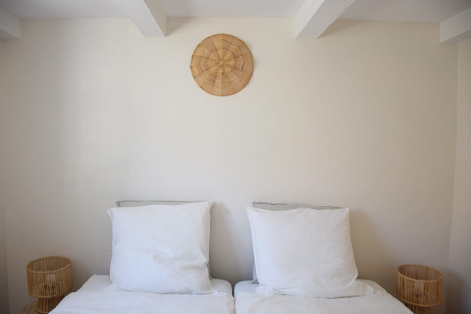 A minimalist bedroom interior with two twin beds and white bedding. Above the beds is a woven decorative piece centered on a plain wall. Two small round wicker bedside tables are placed on either side of the beds.
