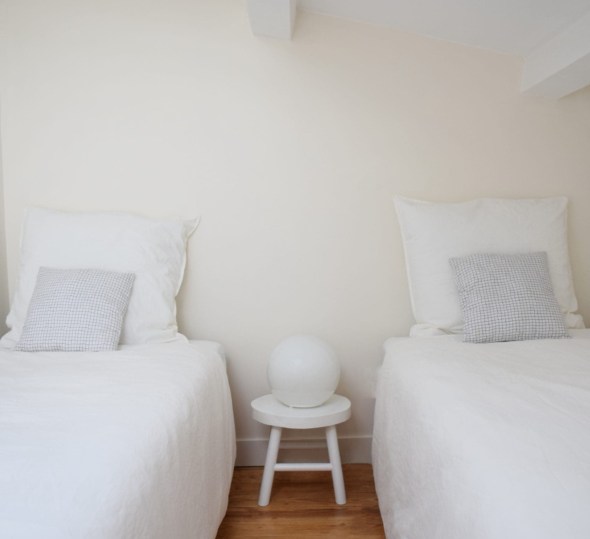 A minimalist room with two white twin beds on either side of a small round white table. Each bed is fitted with a light-gray checked pillow, and the table hosts a white spherical lamp. The walls and bedding are light in color.