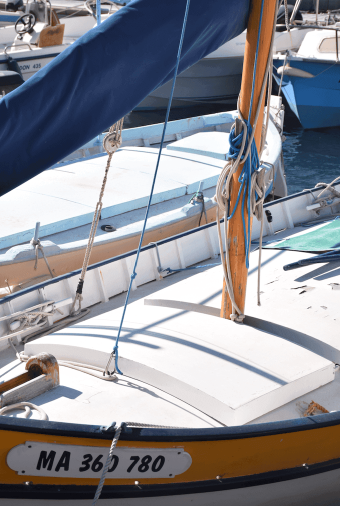 Close-up of a small sailboat with a wooden mast, blue ropes and a partially visible license number “MA 360 780”. The boat is moored in a marina alongside other boats, and the blue water reflects the sunlight.
