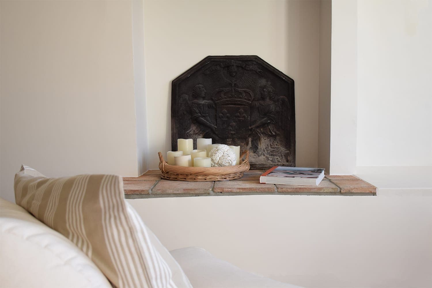 A comfortable living-room scene with a beige striped cushion on a white sofa. Next to it, a shelf houses a wicker tray containing various white candles and a decorative ornament, as well as a closed book. The wall features a dark, ornate mantel cover.