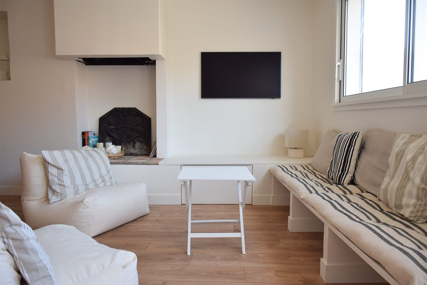 A cozy living room with light wood flooring, wall-mounted TV and fireplace. Seating includes a white armchair and beige benches upholstered with striped cushions. A small white table sits in the center. Natural light enters through a window.