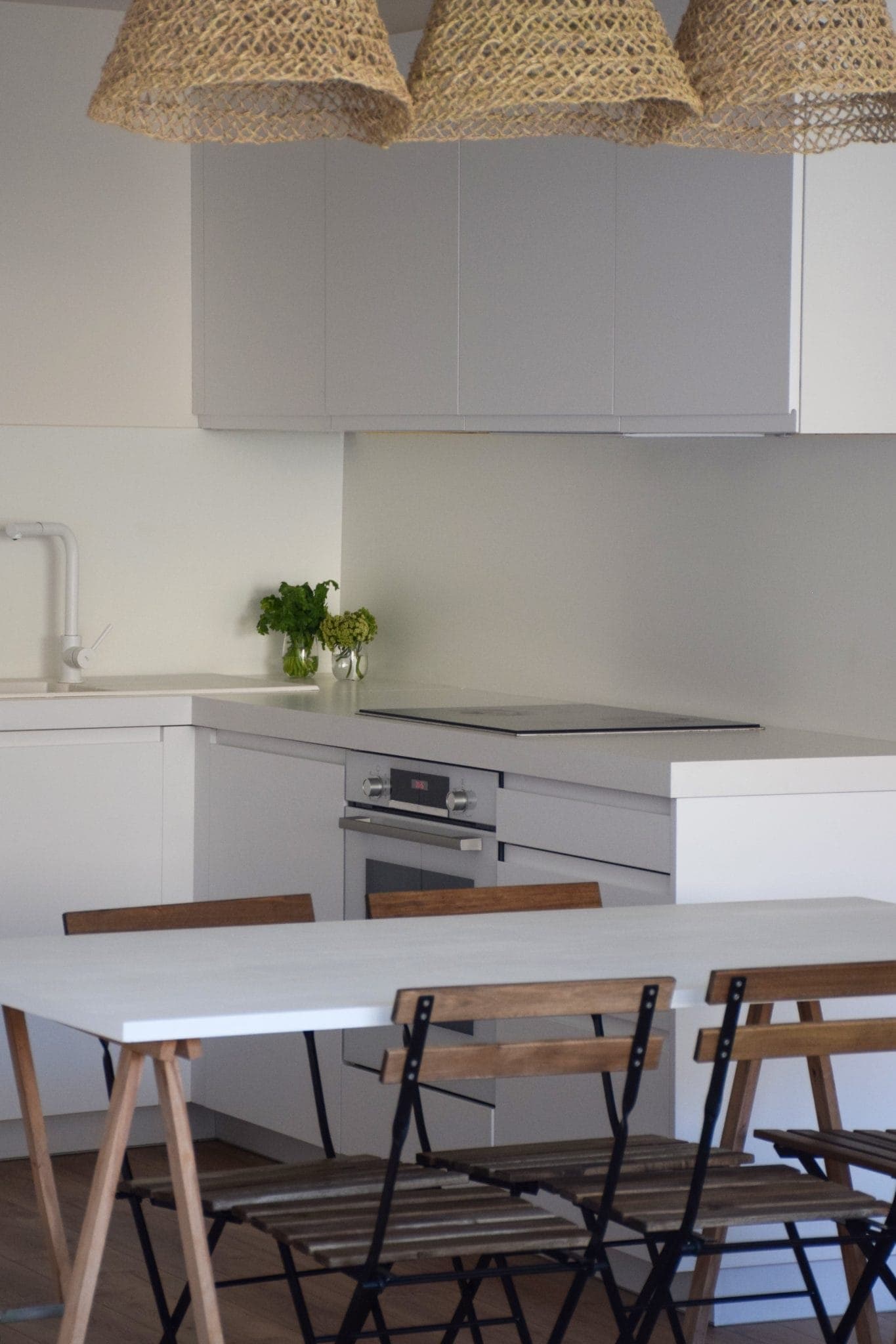 A modern kitchen with white cabinets and built-in oven. A wooden table with folding chairs is in the foreground. Three wicker lights hang above. Fresh herbs in pots sit on the counter, adding a touch of green.