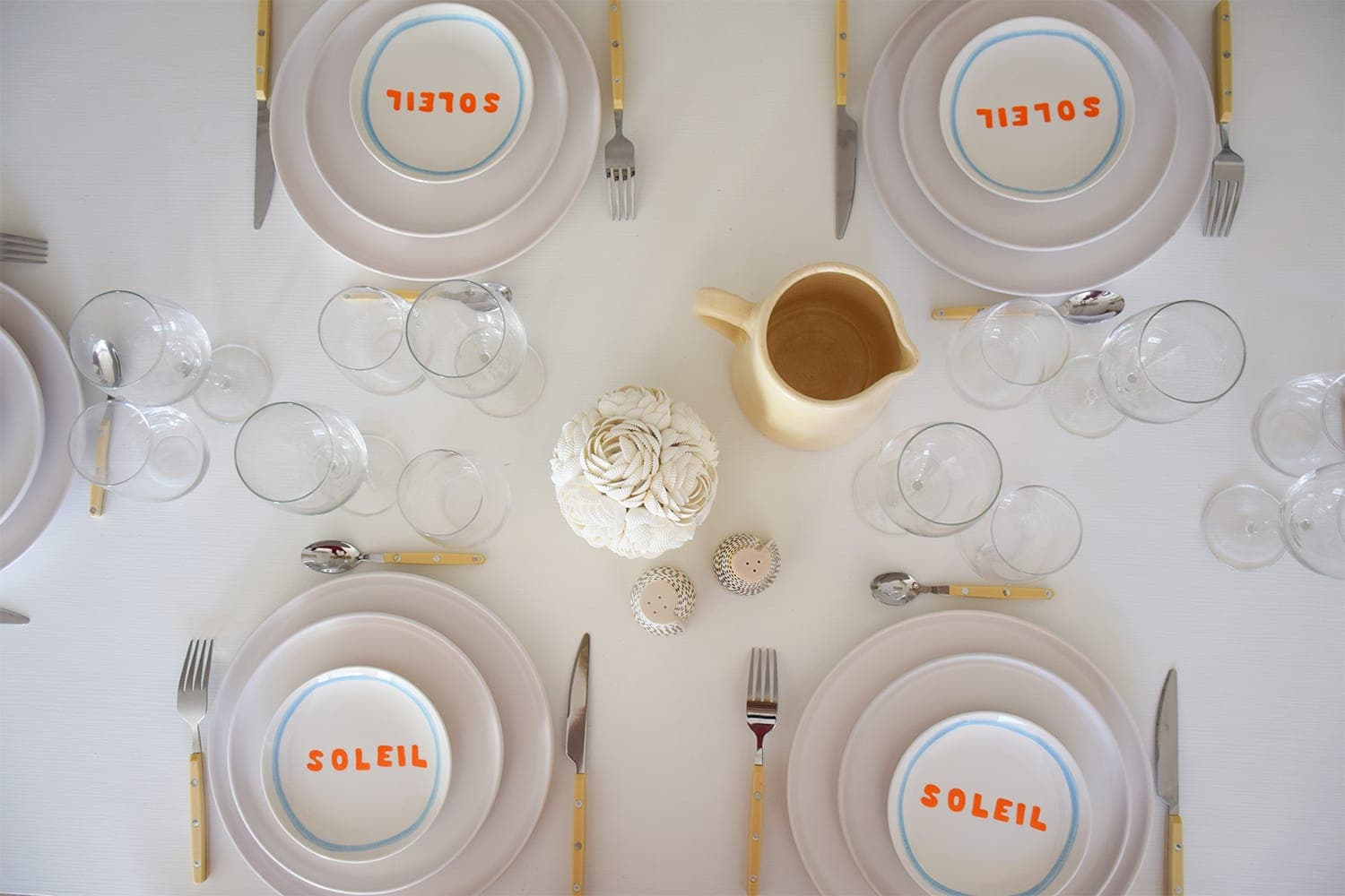 A well-dressed dining table with four place settings, each with a plate labeled “SOLEIL”. The table includes a cream-colored centerpiece, a beige pitcher, several pieces of glassware and a minimalist design on a white tablecloth.