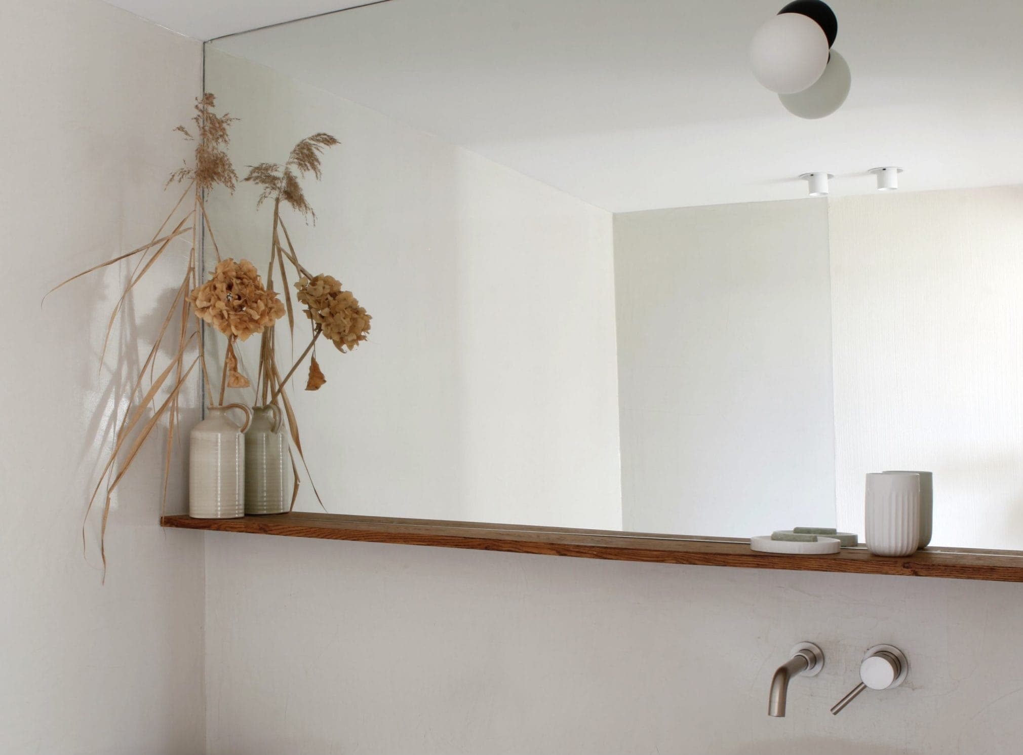 A minimalist bathroom with a large wall mirror, showcasing dried flowers in decorative vases on a wooden shelf. The shelf contains a white mug and a small tray. Two modern wall-mounted faucets are visible under the shelf.