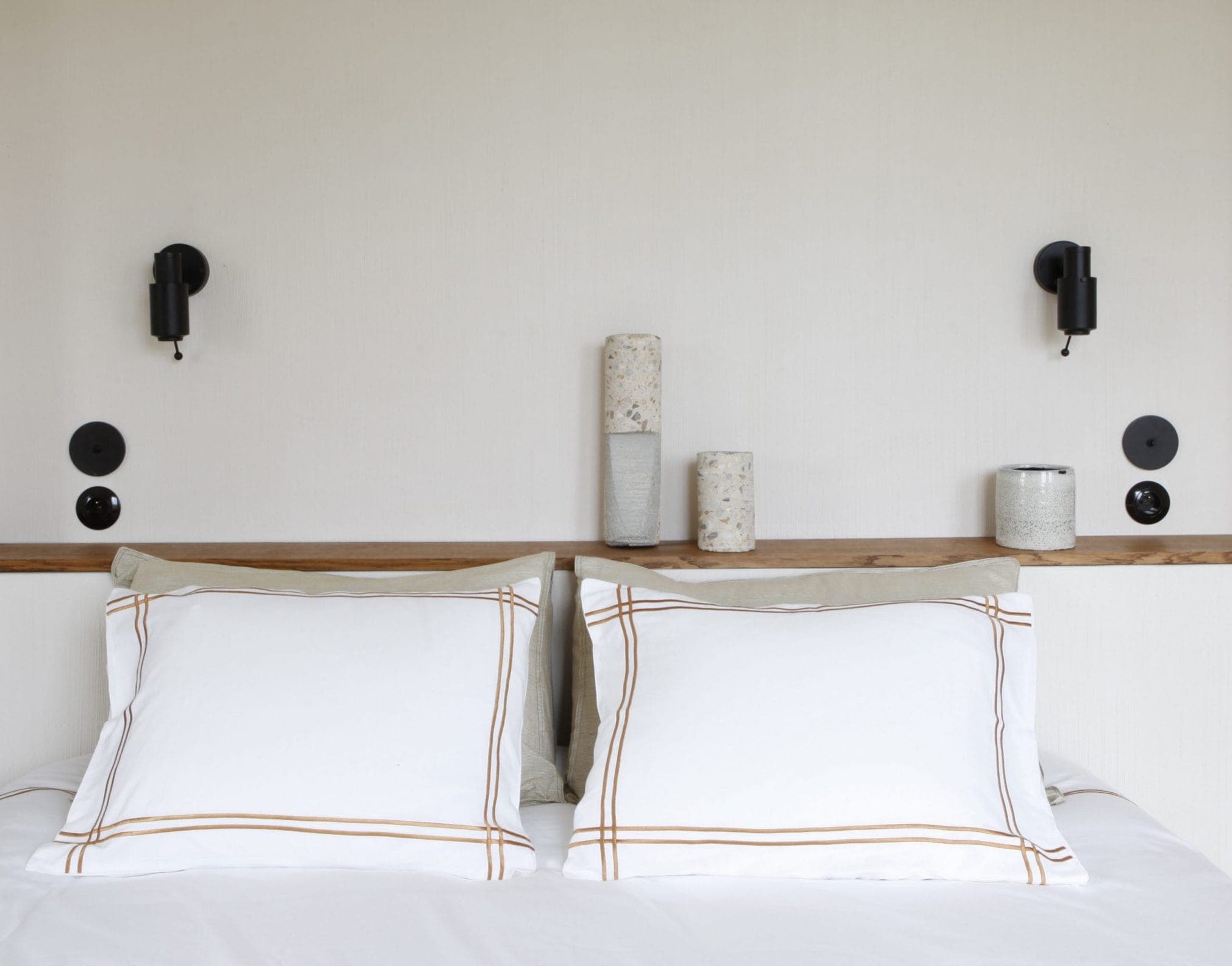 A minimalist bedroom features a well-made bed with white pillows and sheets. The wall behind the bed is adorned with two cylindrical bedside lamps and decorative vases, creating a harmonious, serene atmosphere.