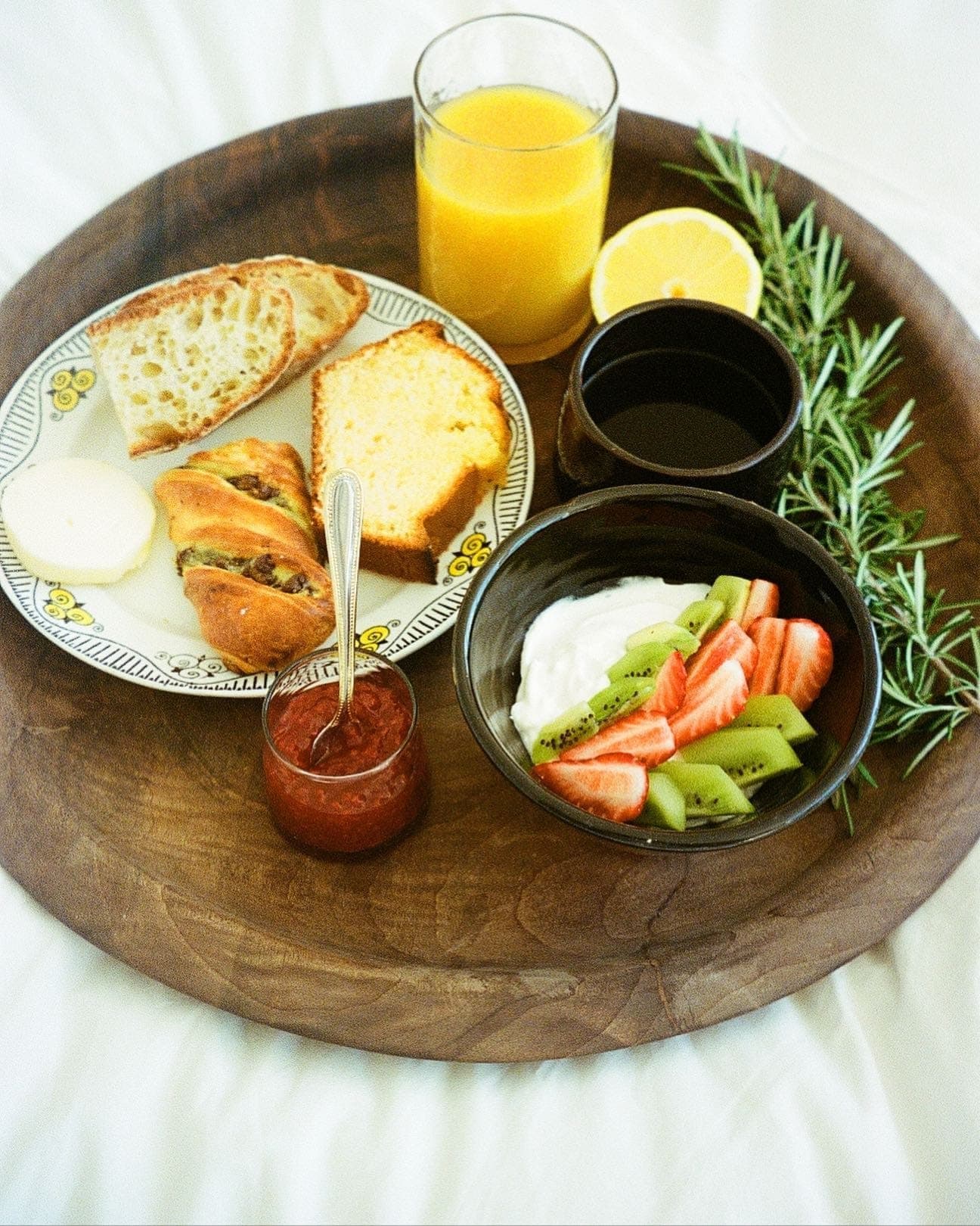 A wooden tray with breakfast items: a glass of orange juice, coffee, half a lemon, sprigs of rosemary, a plate of bread slices, a cake, cheese, a bowl of yoghurt topped with kiwi and strawberries, and a jar of jam with a spoon.