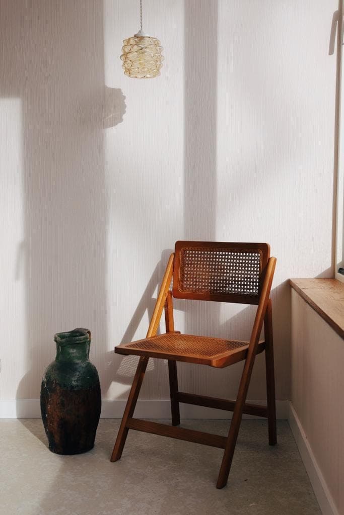 A wooden folding chair with a woven seat and backrest is placed in a sunny corner next to a textured vase. A hanging lamp hangs above, casting soft shadows on the white wall.