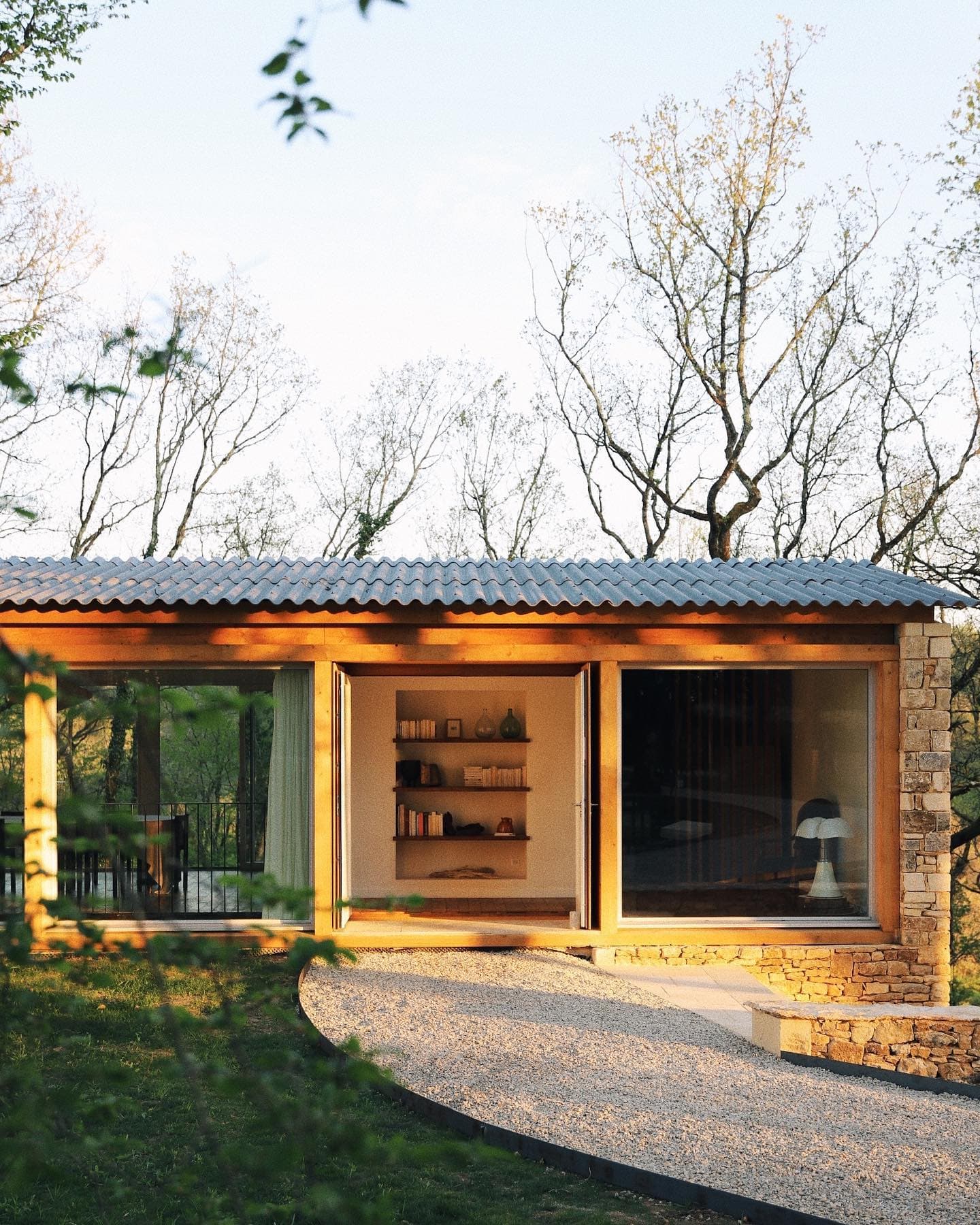 A modern cabin with wood and stone elements. Large windows highlight a warm interior with shelves and decorations. Surrounded by trees and greenery, a curved gravel path leads to the entrance. The setting is peaceful and sunny.