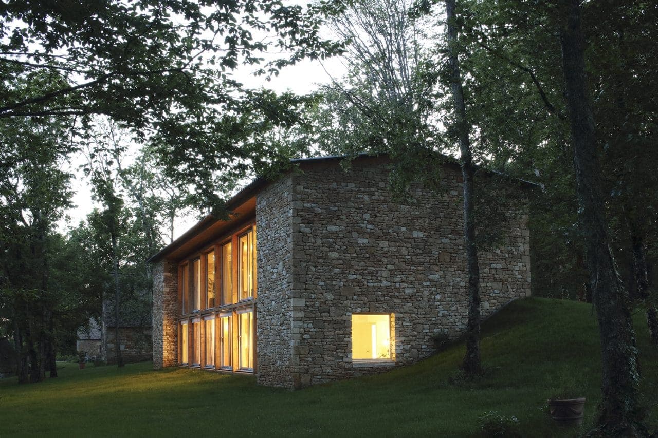 A stone house with large, illuminated windows is nestled among the trees. It stands on a green lawn with a wooded backdrop, suggesting a serene, natural setting. The soft evening light creates a warm ambience in the surroundings.