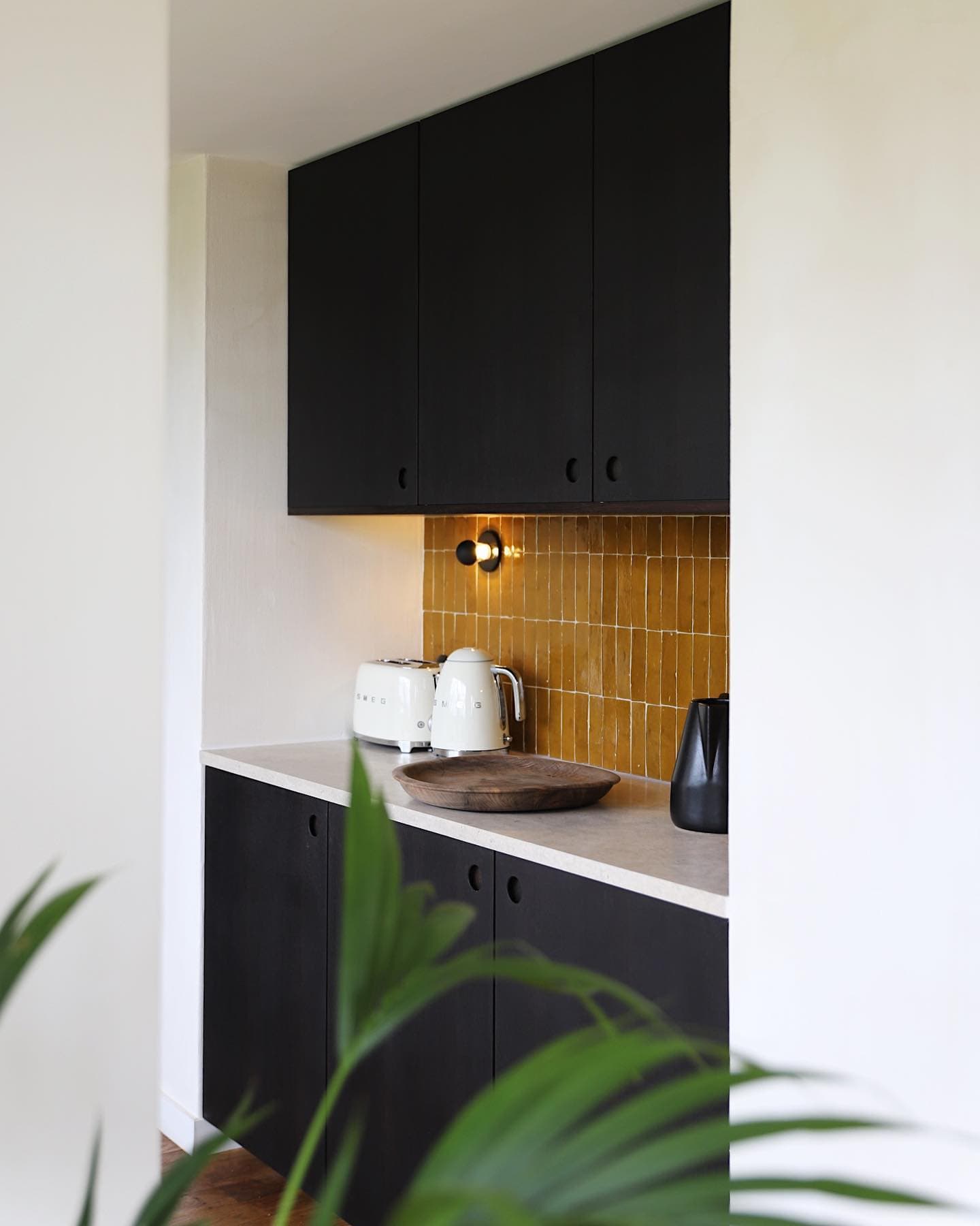 A modern kitchenette with black cabinets, beige countertop and yellow tile backsplash. There are small appliances such as a kettle and toaster on the countertop. A green plant is partially visible in the foreground.
