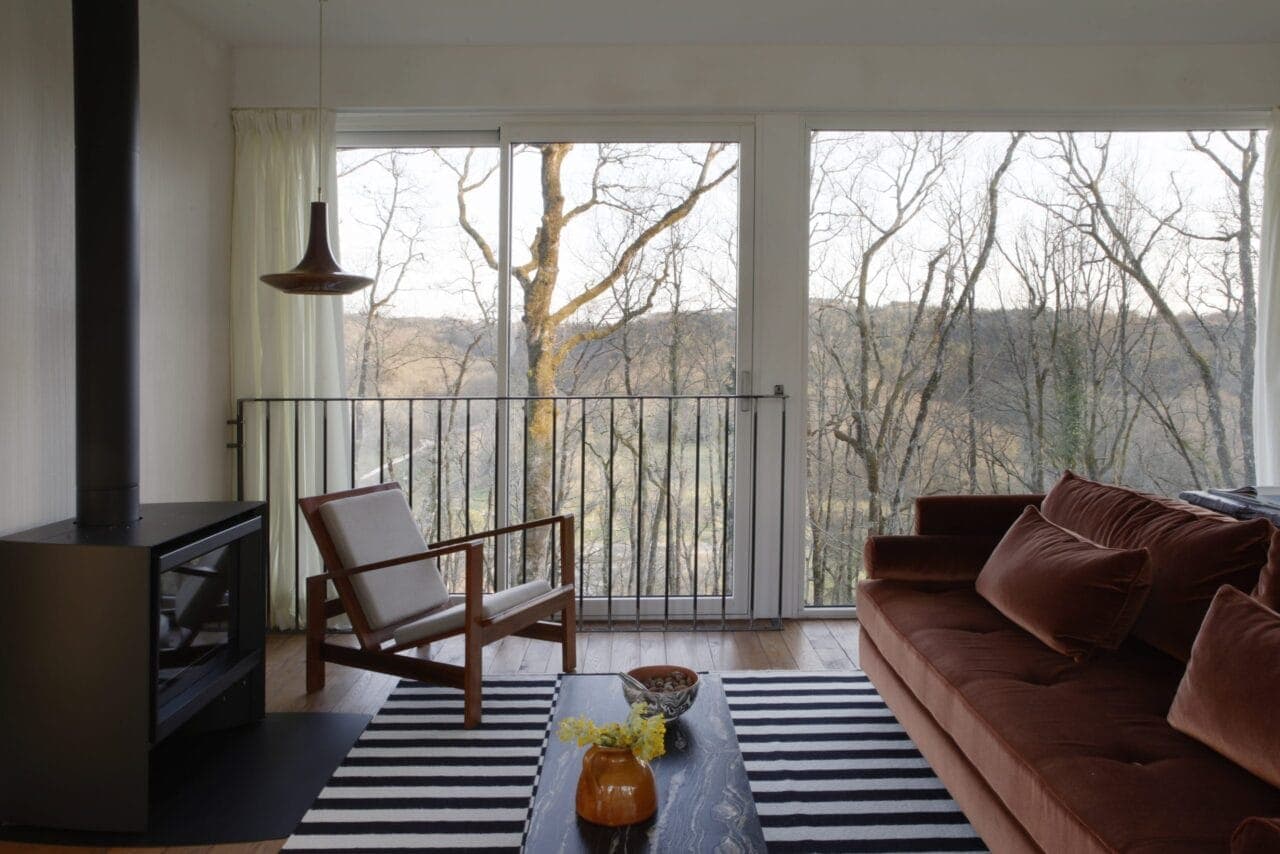 A comfortable living room of modern design, with a brown sofa and wooden chair. A black wood-burning stove sits next to a large window offering panoramic views of leafless trees and rolling hills. A striped rug and a small black table complete the space.