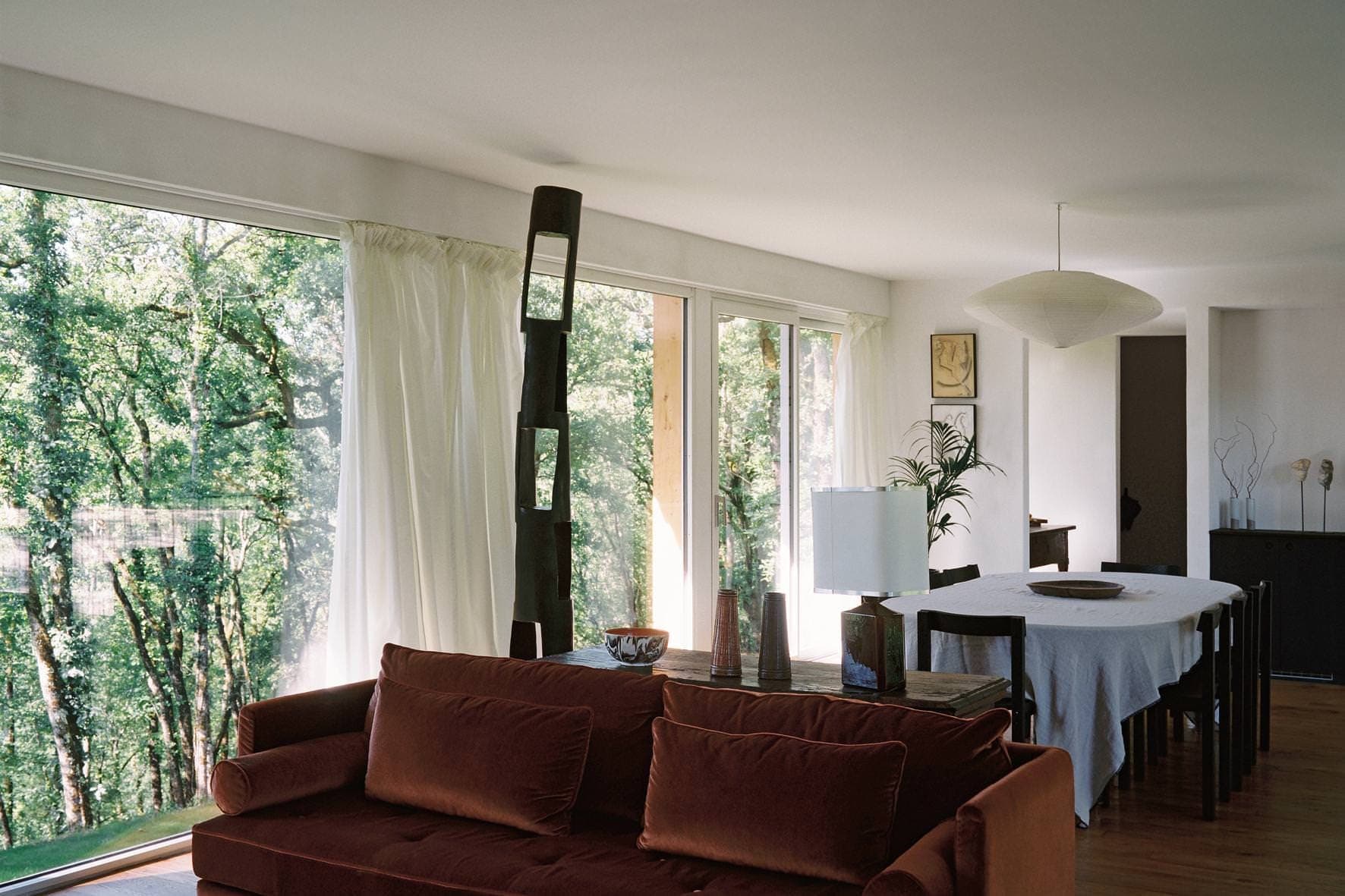 A spacious living room with large windows offering a view of the forest. A rust-colored sofa faces a wooden dining table with a white tablecloth. Various decorative objects and a large lamp are present, as well as a potted plant in the corner.