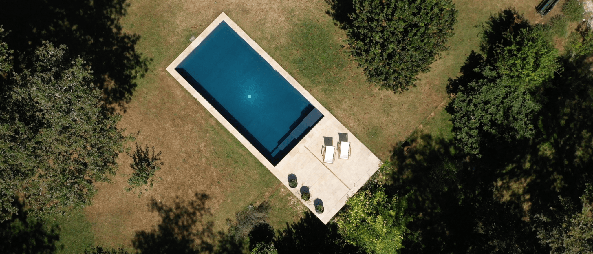 Aerial view of a rectangular pool surrounded by grass and trees. Two deckchairs and three round tables are arranged on the poolside patio.