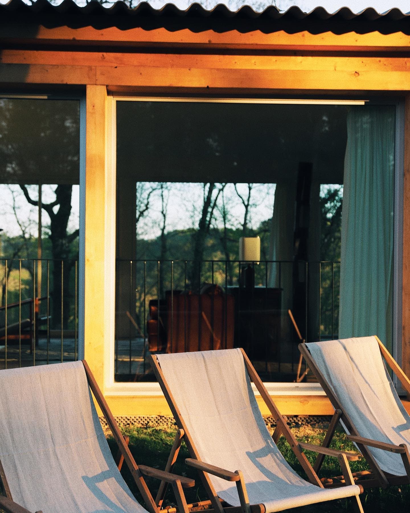 Sun-drenched wooden loungers face a large bay window reflecting the trees. Inside, there's a comfortable interior with a soft-light lamp and sofa. Warm hues suggest a serene late-afternoon ambience.