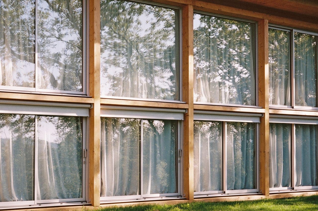 A building facade with large glass windows, each framed by wooden panels. Transparent curtains hang inside. The windows reflect the trees and sky outside. Sunlight casts soft shadows on the grass below.