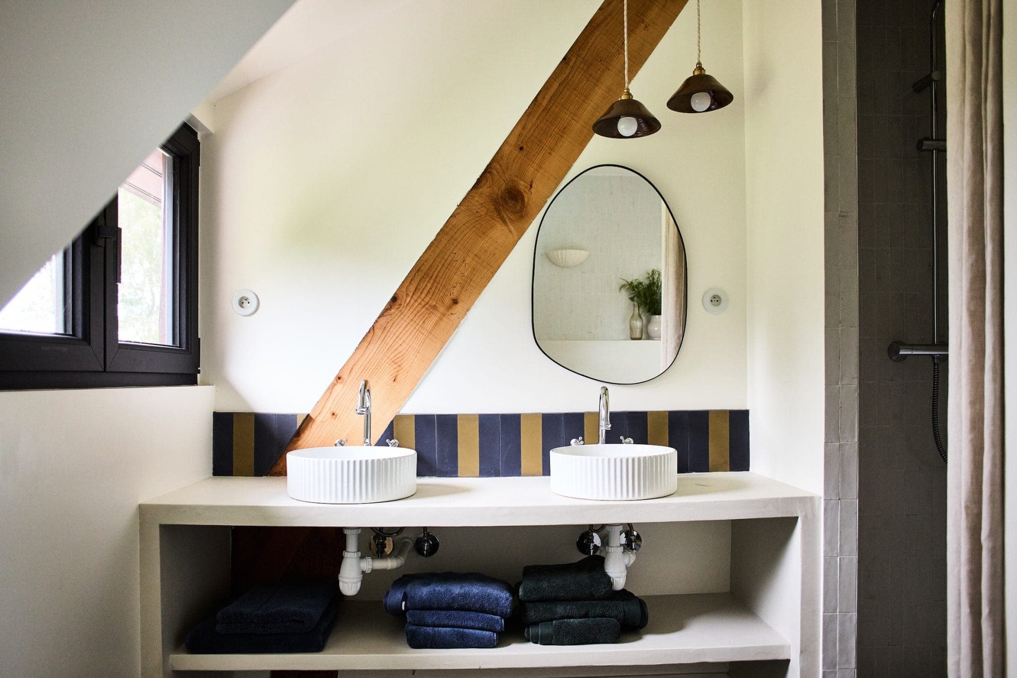 A modern bathroom with a sloping ceiling features two round sinks with a wooden beam. Above the sinks, there's an oval mirror and two pendant lights. Shelves underneath store stacked towels. A window is located on the left, and a shower is partially visible on the right.