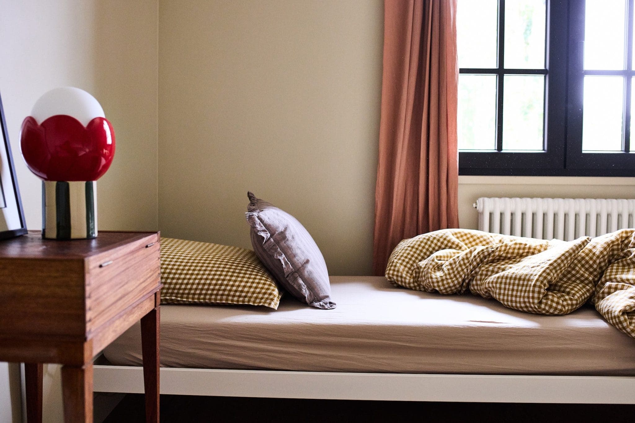 A comfortable room with a single bed against a beige wall. The bed has a brown pillow and a yellow checked comforter. A wooden side table with a red and white lamp stands next to the bed. A window with brown curtains lets in natural light.