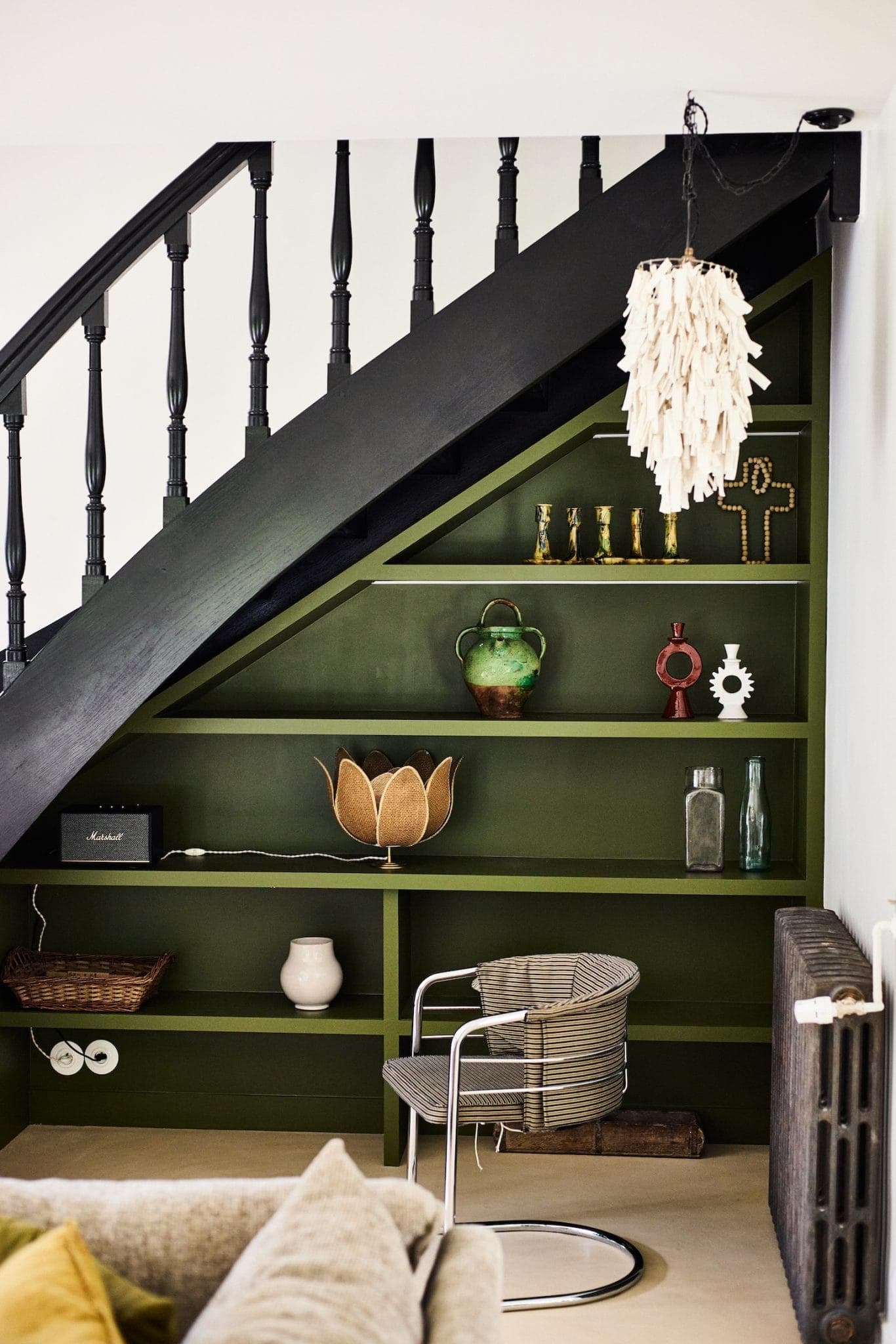A cozy corner with a modern chair, wooden shelves under a staircase and eclectic decorative objects such as vases, a basket and a unique hanging light made of white shells. The shelves are painted dark green.
