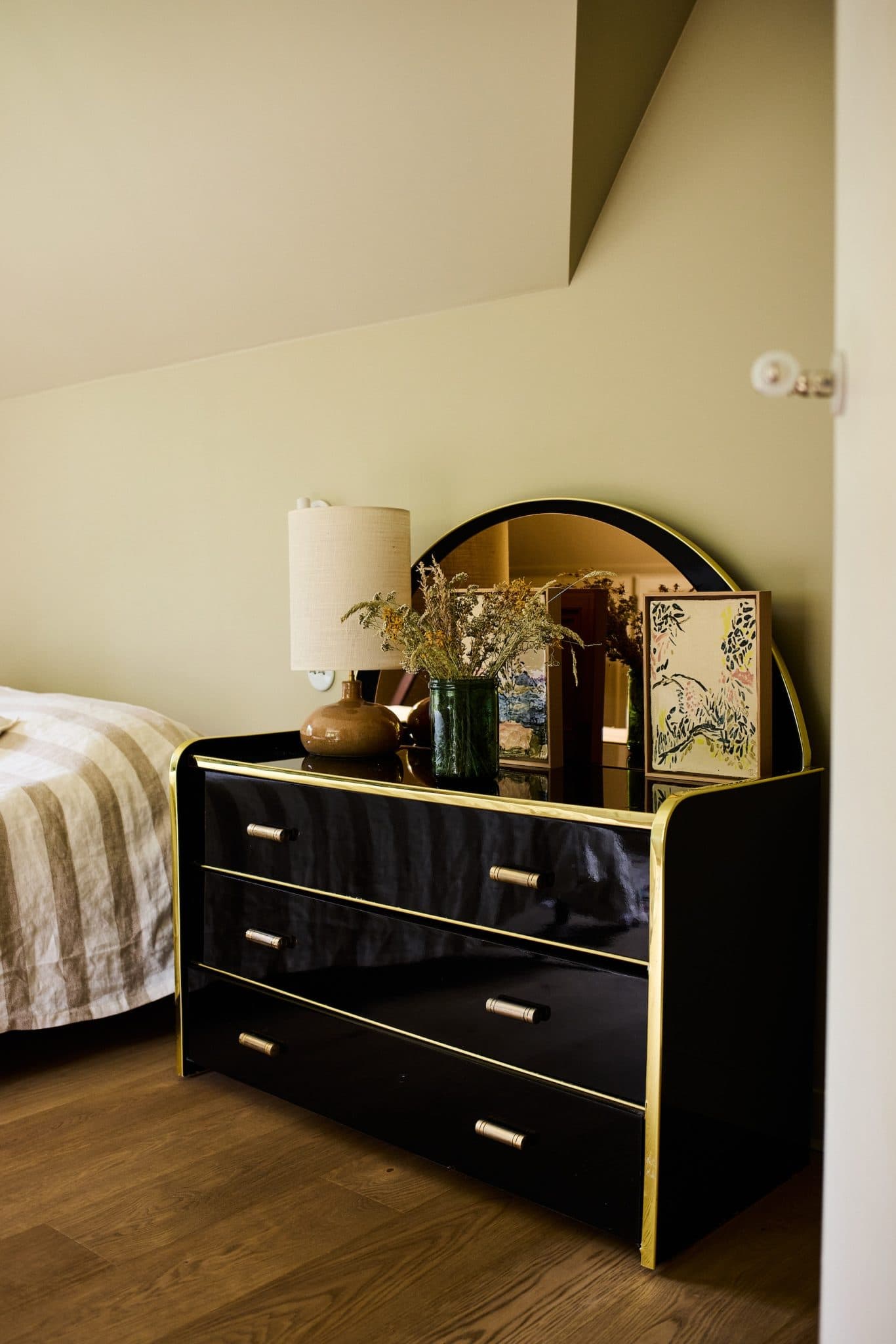 An elegant bedroom corner with a black chest of drawers with gold trim, a round mirror, a lamp, dried flowers in a green vase and a framed decorative painting on top. The wall is light green and part of the bed with a striped blanket is visible.