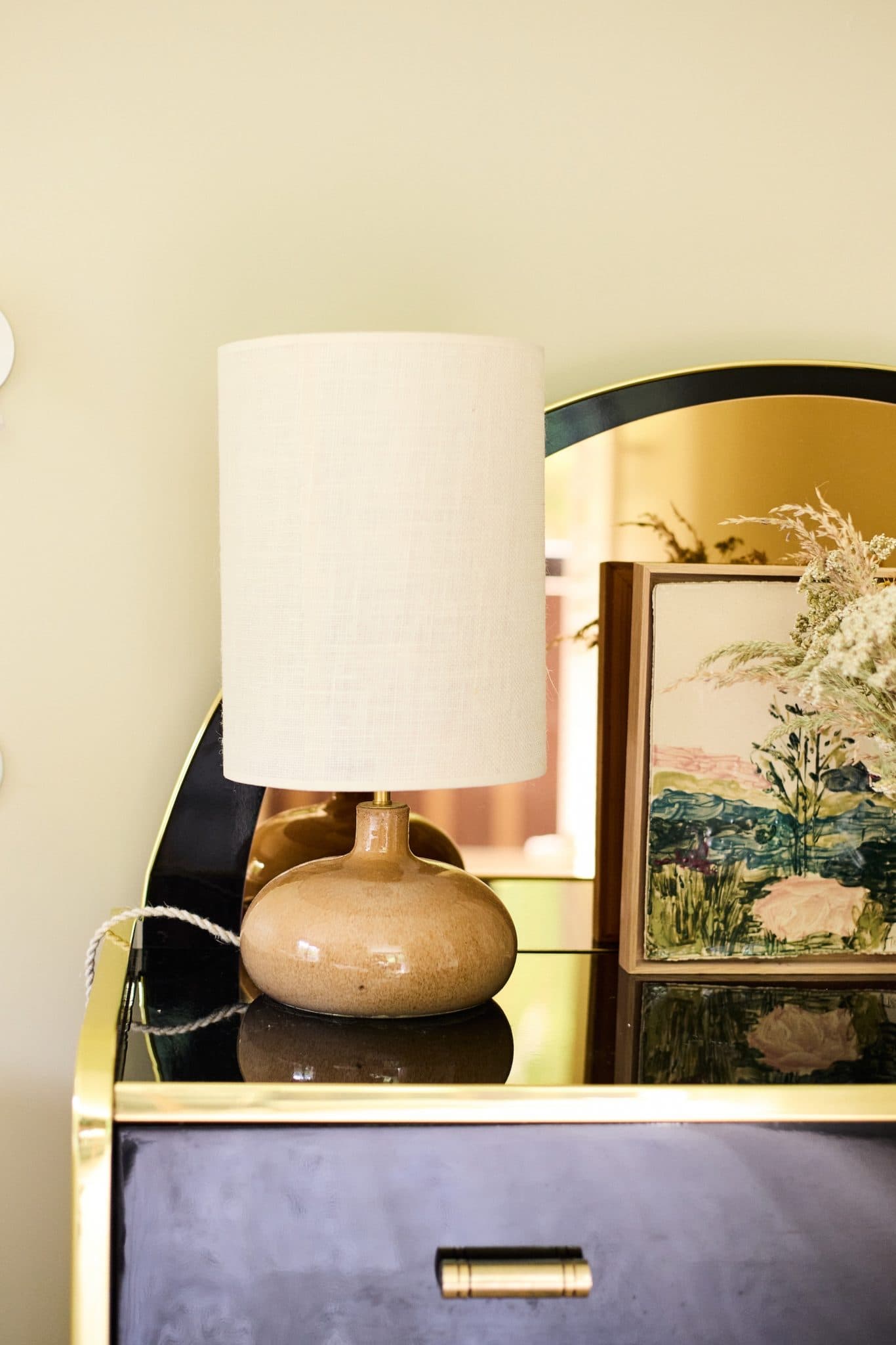 A small beige lamp sits on a shiny black and gold table in front of a curved mirror. Next to the lamp is a framed work of art depicting floral motifs. The back wall is a muted light green.
