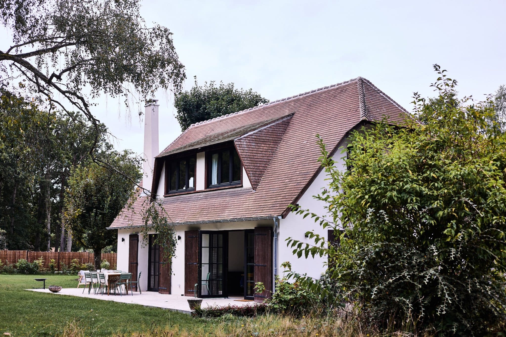 A picturesque cottage with a pitched roof surrounded by lush vegetation. The house features wooden shutters and large windows, as well as a patio with outdoor furniture. Trees and a well-kept lawn complete the serene setting.